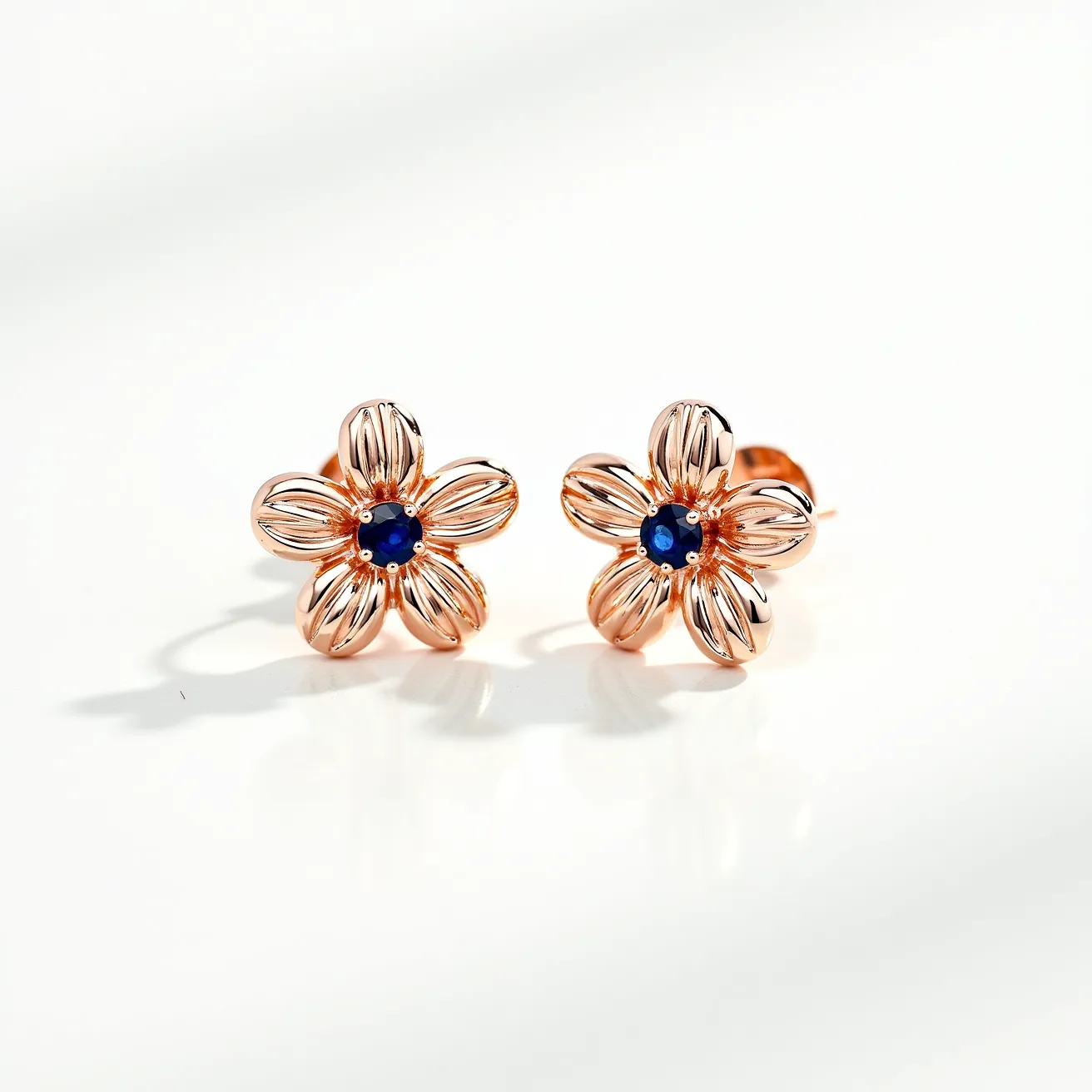 These rose earrings are crafted with a floral design, featuring five intricately shaped petals in a rose gold finish that gives them a sophisticated and elegant appearance. At the center of each earring is a deep blue gemstone, likely a sapphire, cut in a round shape to highlight its vibrant color and clarity. The gemstone is securely held in place by a simple setting that complements the overall design without detracting from the stone's sparkle. The earrings are attached with post and butterfly backs, providing a secure and comfortable fit for everyday wear.