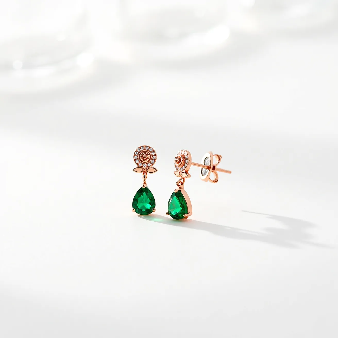 These rose earrings showcase a delicate design with a floral motif at the top, adorned with small, round clear stones set in a circular pattern, resembling petals. The earrings are crafted from a rose gold-colored metal, enhancing their elegant appearance. A pear-shaped green stone, with a brilliant cut, dangles below, set in a classic claw setting that accentuates its vibrant hue. The attachment mechanism features a standard post and butterfly clutch back, ensuring a secure fit when worn. These earrings combine floral and gemstone elements, creating a striking and sophisticated accessory.