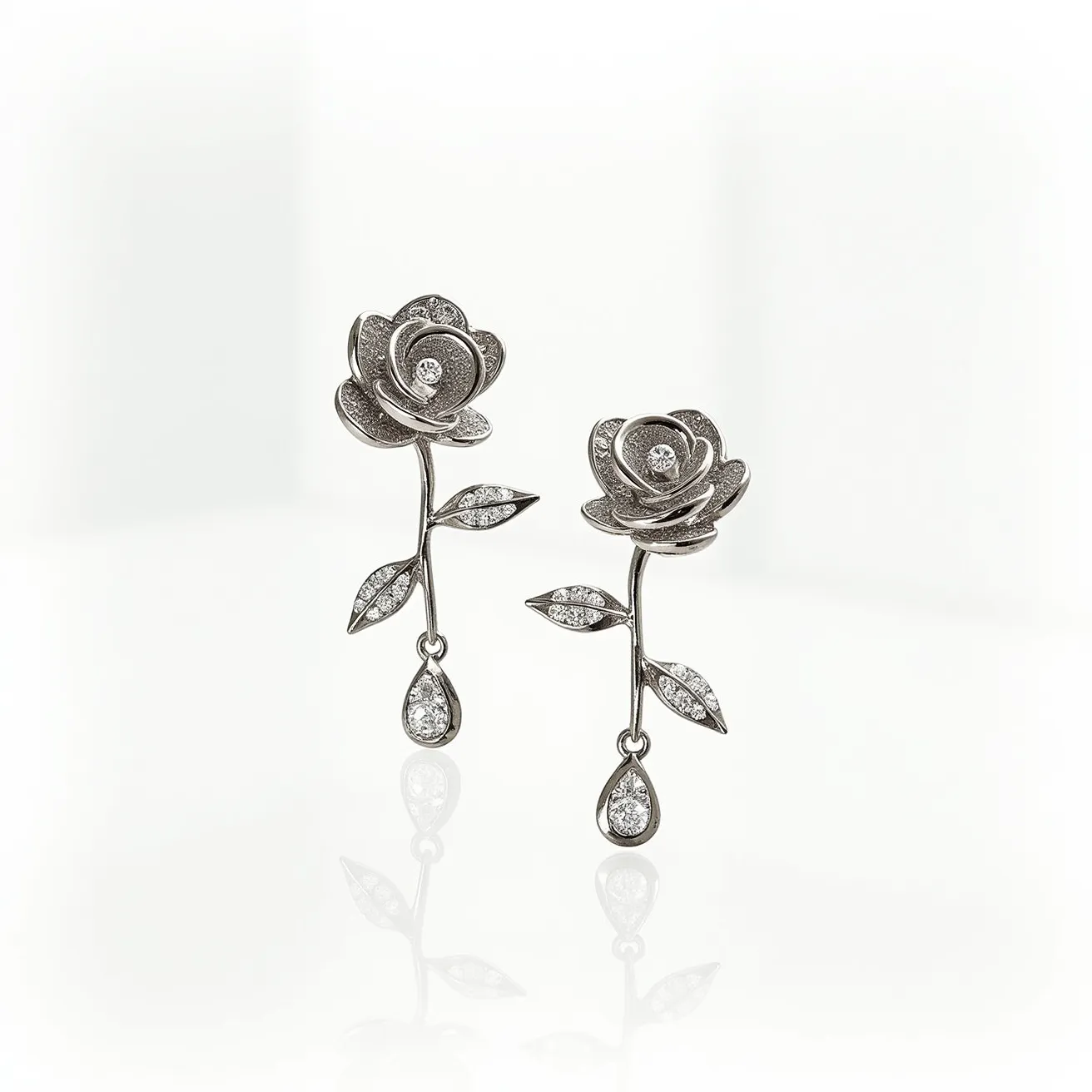 These rose earrings feature a delicate floral design, crafted from what appears to be a polished metal, likely white gold or platinum, which gives it a luxurious sheen. The roses showcase intricate layering of petals, each embedded with small, round-cut diamonds that are prong-set to enhance their sparkle. In the center of each rose, a larger diamond acts as a striking focal point. The stem of the earrings is adorned with leaf-shaped elements, each studded with tiny diamonds, continuing the motif with elegance. At the end of the stem, a teardrop-shaped pendant dangles, set with pave diamonds for added brilliance. The earrings seem to utilize a post or hook attachment, suitable for pierced ears.

