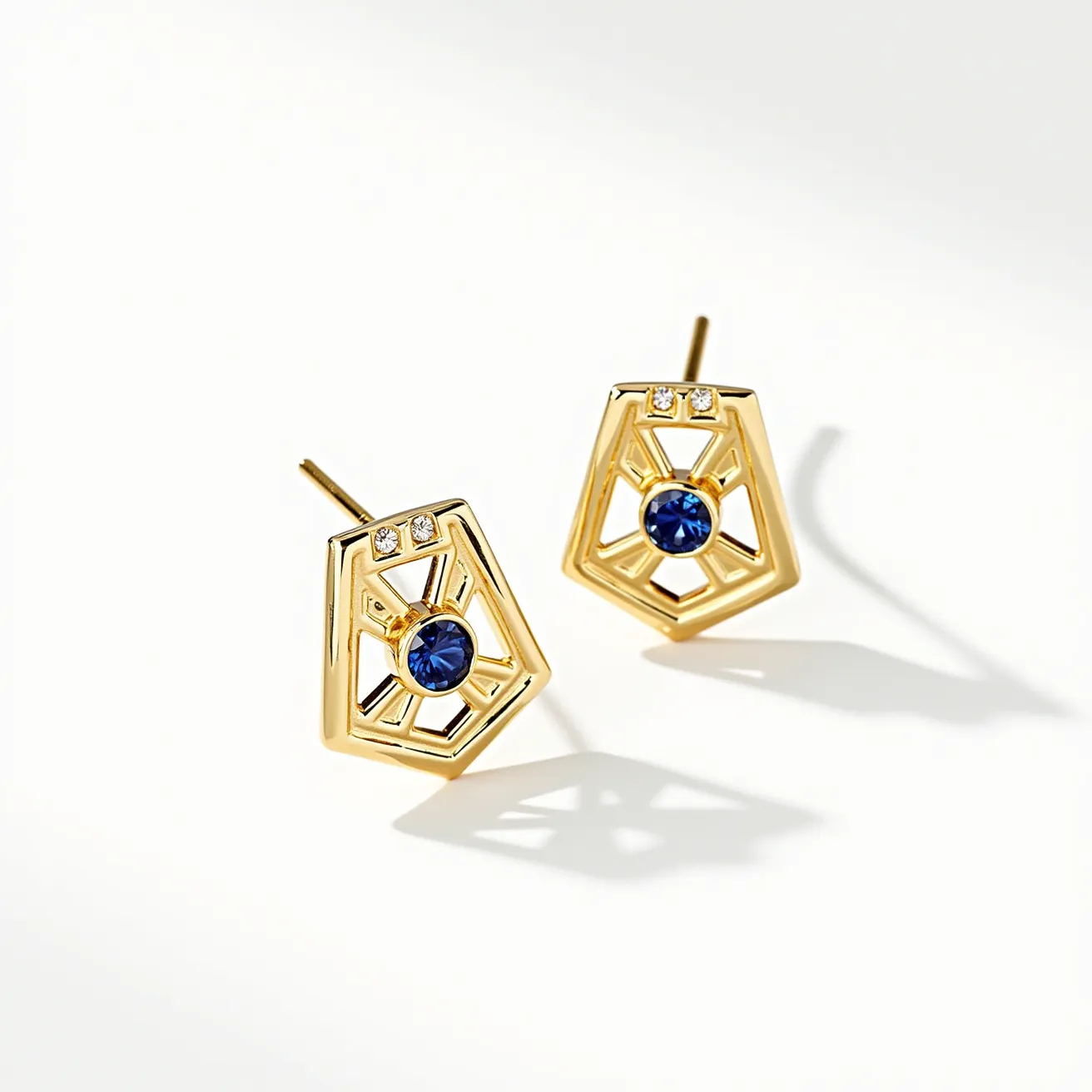 These sapphire earrings showcase a striking design featuring deep blue round-cut sapphires encased in a geometric gold setting. The sapphires are centrally placed, surrounded by a gold framework with an intricate openwork pattern that adds an airy elegance to the overall appearance. Each earring is adorned with two small, round-cut clear stones at the top, likely diamonds, enhancing their luxurious appeal. The earrings use a traditional post backing, ensuring a secure and comfortable fit on the ear.