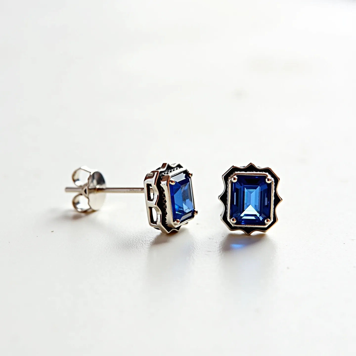 These sapphire earrings feature a striking pair of rectangular blue sapphire stones, each carefully cut in an elegant emerald style to showcase their depth and color. The sapphires are securely set in a stylish silver prong setting that incorporates a scalloped border design, enhancing their classic appeal. The stud earrings are equipped with a standard post-and-clutch backing system, ensuring they remain comfortably and securely fastened when worn. The combination of the deep blue sapphires and the polished silver setting creates a timeless and versatile accessory suitable for various occasions.