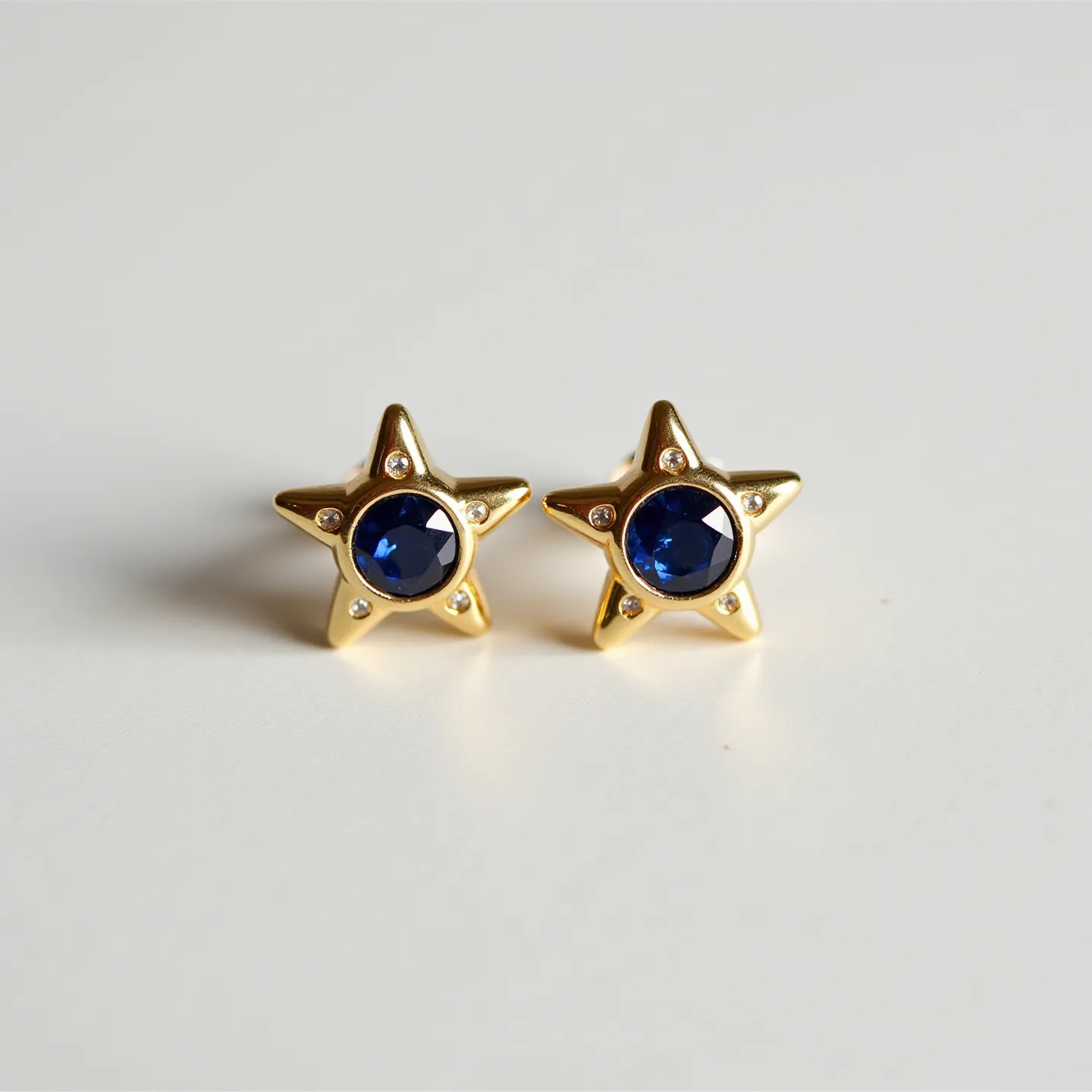 These sapphire earrings feature a striking design with star-shaped settings crafted from a gold material. Each earring prominently showcases a deep blue, round-cut sapphire at the center, adding a rich color contrast. The sapphires are securely bezel-set within the star, which includes small, clear stones embedded at each of the five points, enhancing the overall sparkle. The earrings likely use a classic post backing for secure attachment, blending functionality with elegant design.