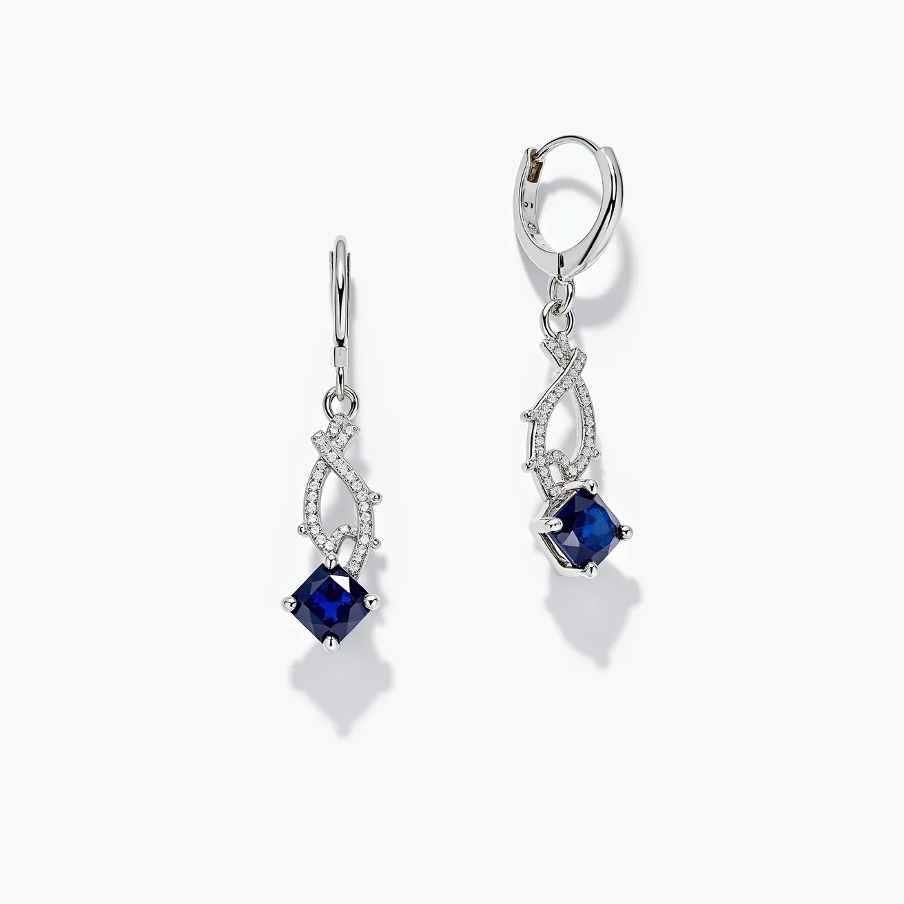 These sapphire earrings feature striking blue sapphires cut in a square shape, skillfully held in a prong setting. The sapphires are further enhanced by a delicate arrangement of smaller, presumably diamond stones that are embedded in a whimsical, interwoven design above the main gem. The intricate pattern adds a touch of elegance and sophistication to the overall appearance. These elements are all set in a polished, likely silver or white gold metal, which complements the rich hue of the sapphires. The earrings are equipped with a lever-back clasp, ensuring a secure and comfortable fit when worn.