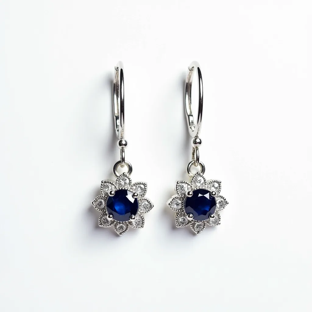 These silver dangle earrings feature a striking design, showcasing a central dark blue gemstone, likely a sapphire, in each earring. The stones are cut in a round shape and are secured within a classic prong setting, surrounded by a decorative floral motif crafted from silver, which adds an intricate touch to the overall look. The earrings are attached to the ear with a hoop-style clasp, ensuring both elegance and security when worn. The polished silver complements the deep hue of the gemstones, creating a sophisticated and timeless appeal.