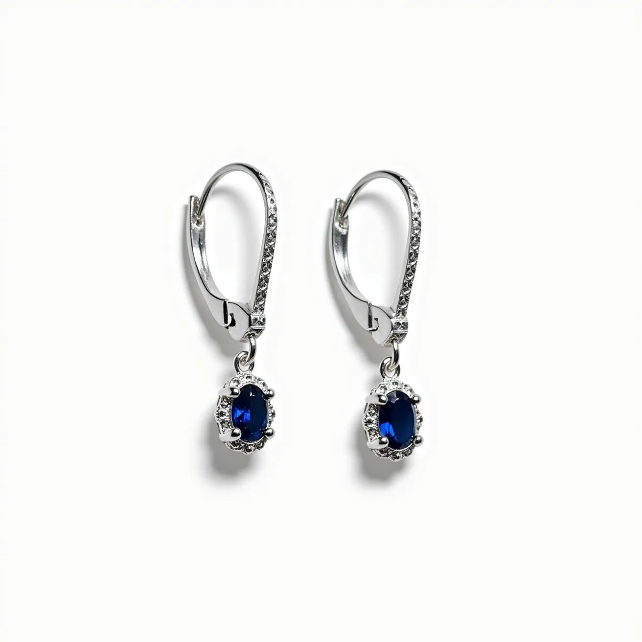 These silver dangle earrings feature an elegant design with a pair of striking oval blue stones set at the center, possibly sapphires, which are surrounded by a decorative halo of smaller clear stones in a prong setting. The earrings are crafted from a polished silver metal, which adds a sleek and modern finish. The attachment includes a secure lever-back clasp, ensuring both comfort and security when worn. The combination of deep blue hues and sparkling accents creates an eye-catching contrast that enhances the overall aesthetic of the earrings.