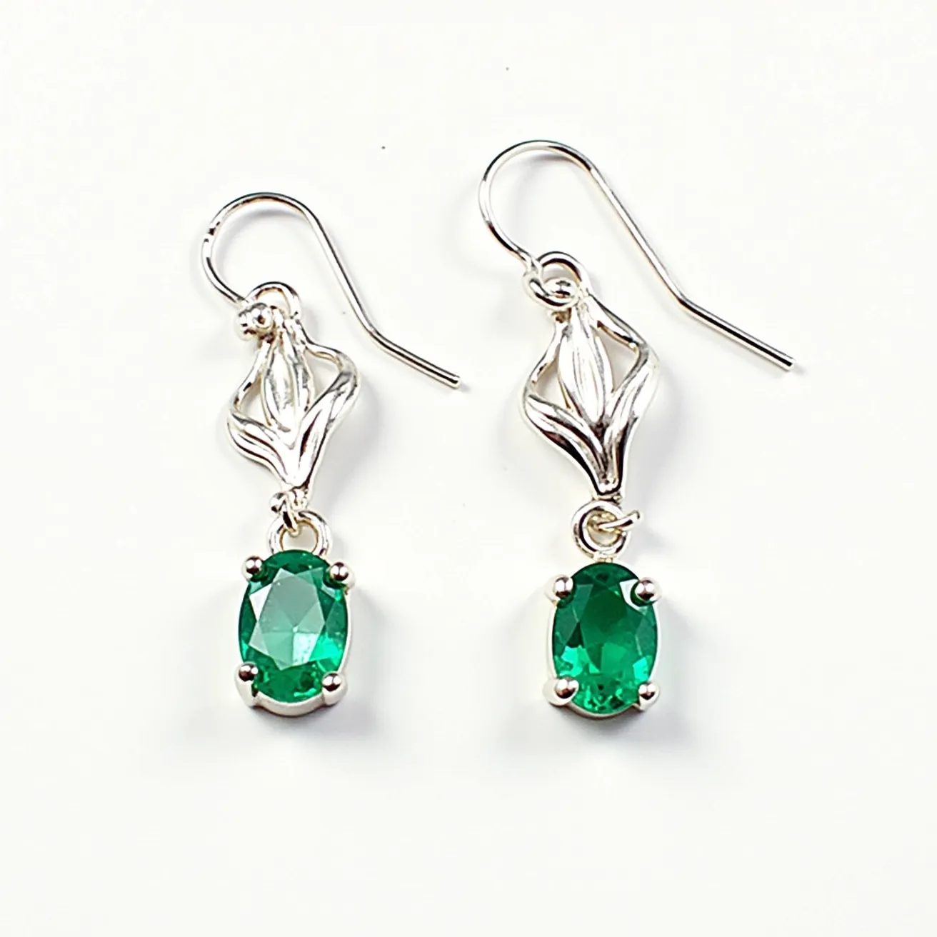 These silver dangle earrings feature a delicate filigree design crafted from polished silver, enhancing their elegance. Set within each earring is a vibrant green oval gem, likely an emerald or similarly colored gemstone, showcasing a faceted cut that catches the light beautifully. The stones are secured in a prong setting, which provides both security and visibility to the gems' brilliance. These earrings utilize a classic hook attachment, allowing them to dangle gracefully when worn, adding a touch of sophistication to any ensemble.