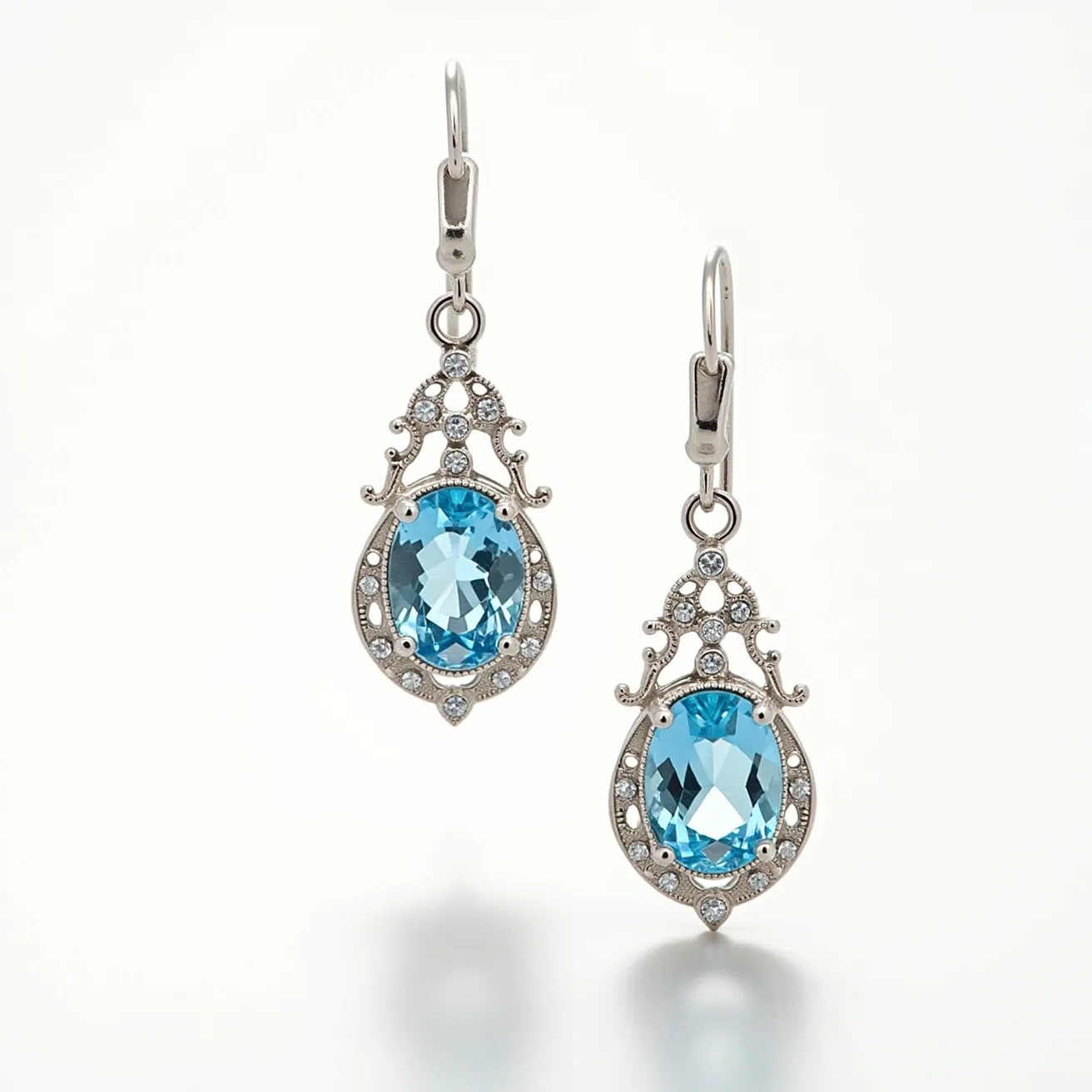 These silver drop earrings are elegantly crafted with intricate metalwork, featuring an ornate design that beautifully frames a large, oval-cut blue gemstone at the center of each earring. The stones are securely set in a bezel setting, surrounded by smaller clear gems that enhance their sparkle and appeal. The earrings are designed with a hook clasp attachment, ensuring both security and ease of wear. The combination of the shimmering silver and the captivating blue stones creates a striking contrast, resulting in a sophisticated and timeless accessory.