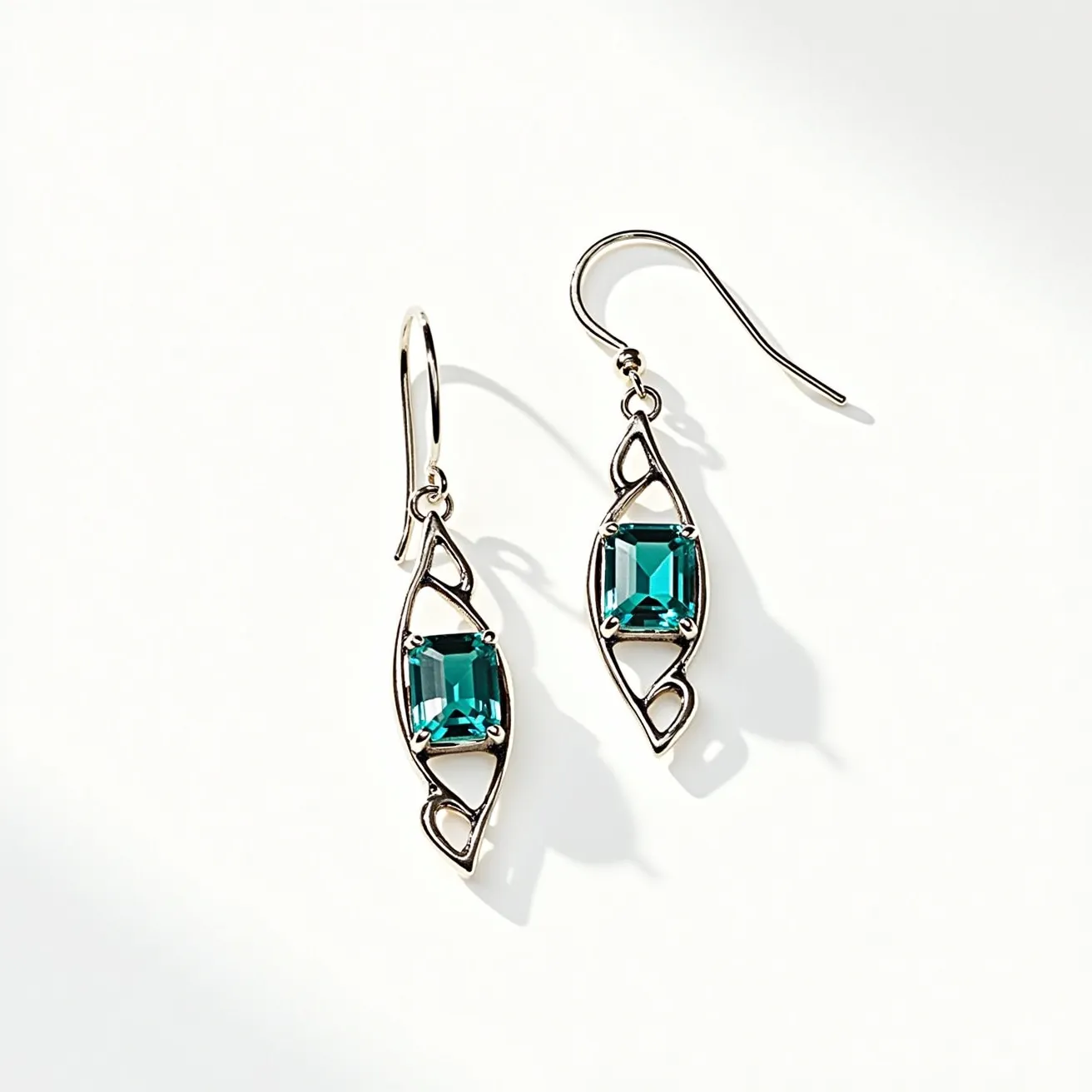 These silver drop earrings feature an elegant and intricate design with a sleek, shiny metal structure that frames the centerpiece. Each earring holds a stunning square-shaped blue gemstone, secured by a prong setting that highlights its vibrant color and cut. The gemstones are likely a teal blue, adding a striking contrast to the silver framework. The earrings are designed with a simple yet effective hook clasp, which ensures ease of wearing and adds to their graceful drape. The overall craftsmanship suggests a blend of contemporary style and classic elegance, making these earrings a versatile accessory for various occasions.