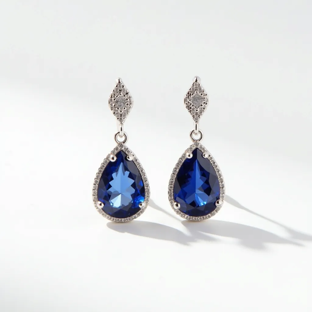 These silver drop earrings feature a sophisticated design with pear-shaped blue gemstones, likely sapphires, that are set in a teardrop silver frame. The gemstones exhibit a faceted cut that enhances their deep blue hue and sparkle. Surrounding each blue stone is a border of small, round white stones, possibly diamonds or cubic zirconia, providing a striking contrast and additional brilliance. The earrings are connected by a small linking loop to a marquise-shaped upper section, adorned with additional white stones. This elegant design is completed with a post or stud backing that offers secure attachment to the ear.