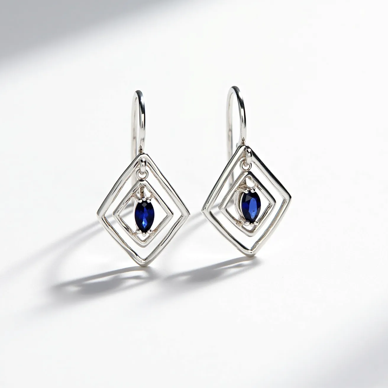 These silver earrings feature a modern geometric design with overlapping rhombus shapes crafted from polished silver. At the center of each earring, a deep blue oval gemstone is set in a secure four-prong setting, adding a vibrant contrast against the silver framework. The gemstones appear to be faceted, enhancing their sparkle and allure. The earrings are designed with a simple hook clasp, allowing for easy and secure wear.