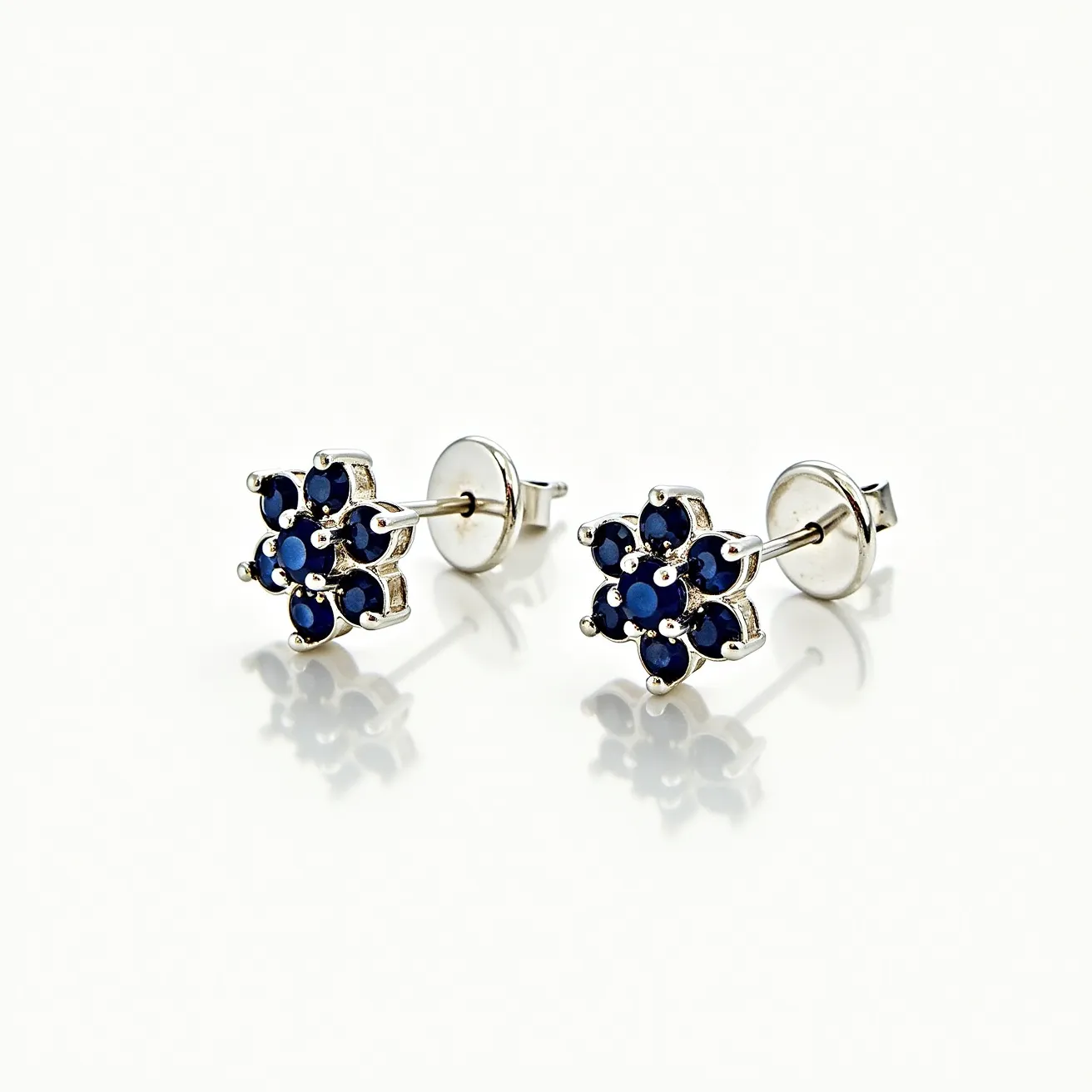 These silver earrings feature a floral design, each with six small round dark blue gems arranged in a circular shape around a central gem. The gems appear to be set using prongs, offering a secure hold while highlighting their sparkling facets. The earrings are crafted from silver, enhancing the vibrant dark blue color of the stones. They are equipped with a simple post and butterfly clasp for secure attachment to the ears, making them both elegant and practical for daily wear.