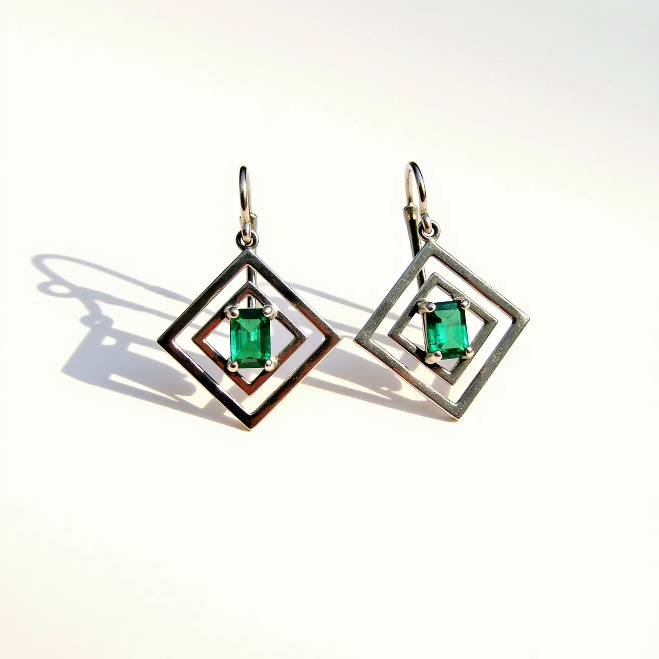 These silver earrings feature a geometric design with two overlapping square frames, creating an intriguing layered effect. At the center of this design is a striking green gemstone, likely an emerald, cut in an emerald shape and securely set with four prongs that highlight its vivid color. The earrings are attached with simple, elegant hooks, allowing for easy wear and movement. The combination of the cool, polished silver and the vibrant green gemstone gives these earrings a modern and sophisticated look.