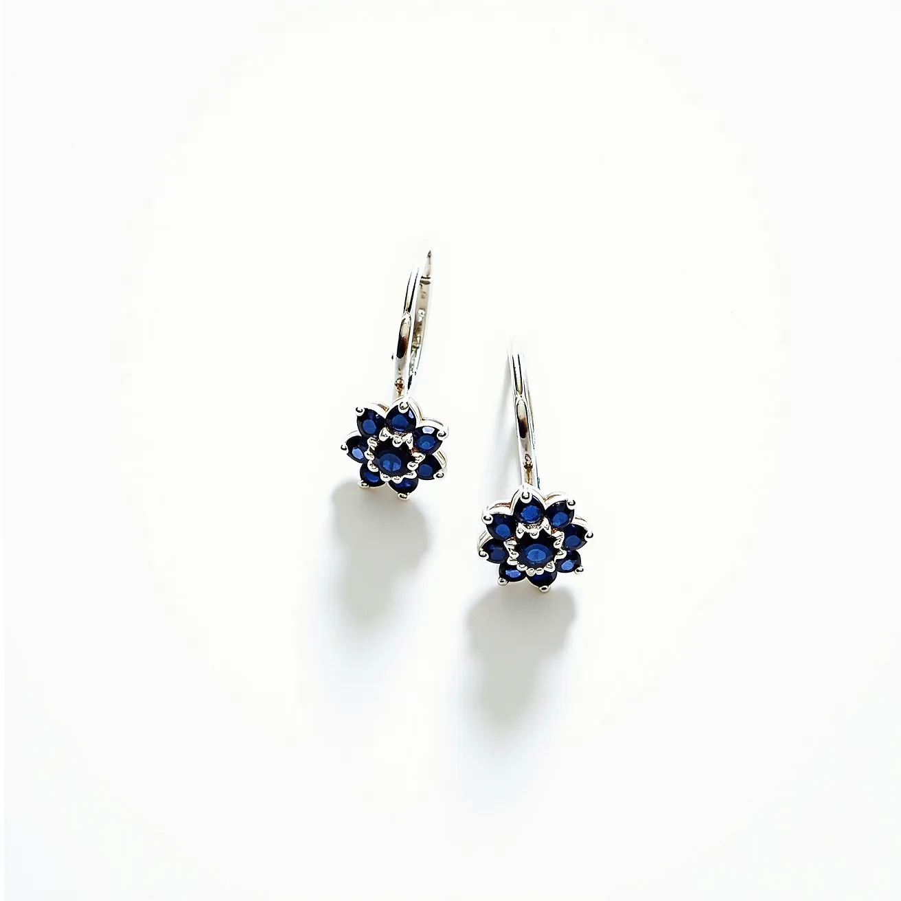 These silver earrings feature an elegant floral design made up of blue gemstones. The gemstones appear to be round-cut sapphires, arranged beautifully in a prong setting that enhances their vivid color and sparkle. The earring design showcases a leverback clasp, ensuring secure wear while adding a touch of sophistication. The silver metal complements the gemstones, creating a striking and harmonious appearance.