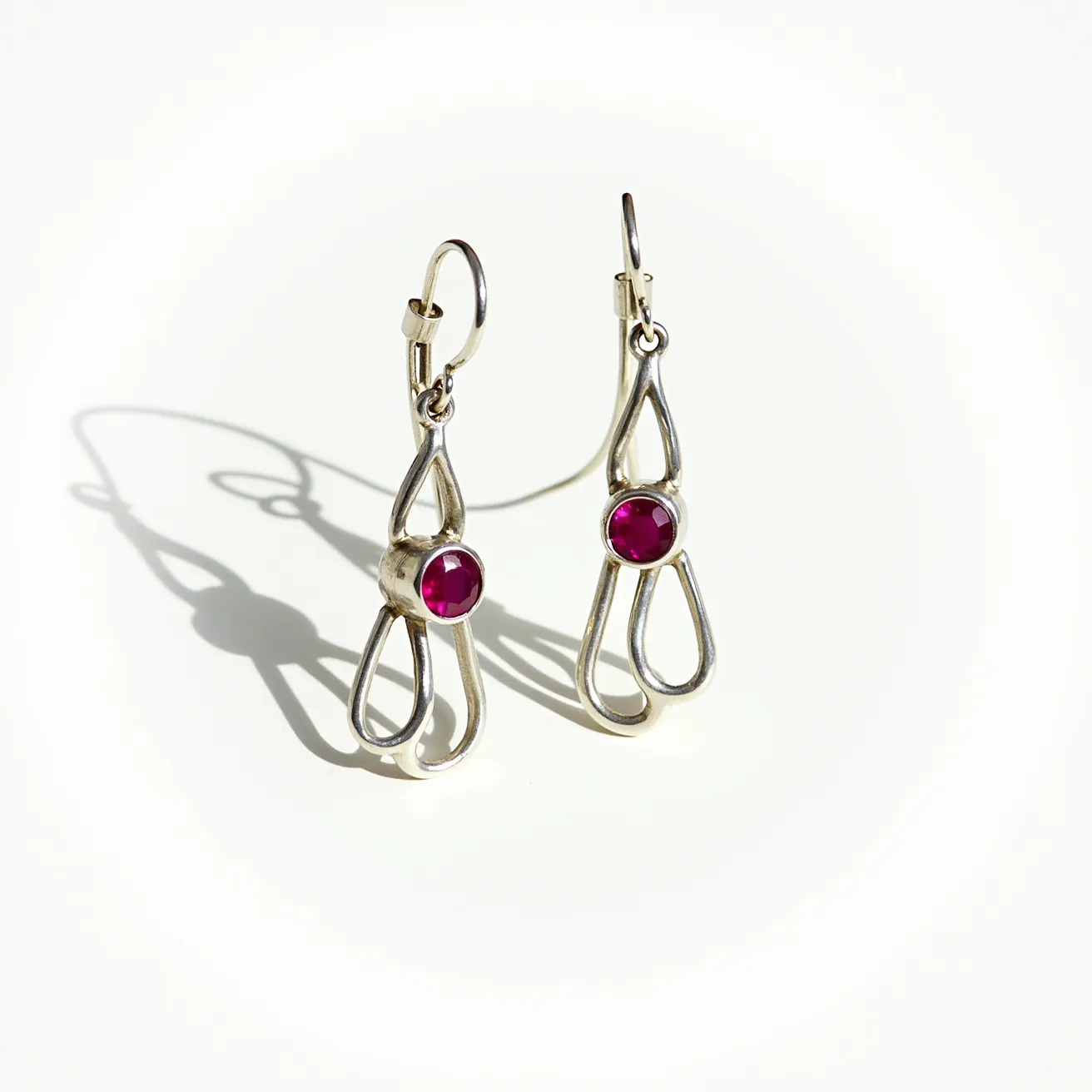 These silver earrings feature an elegant drop design crafted from polished metal. Each earring showcases a round, deep red gemstone set in a bezel setting, which adds a vibrant pop of color and highlights the stone's smooth surface and rich hue. The open framework of the earrings enhances their airy appearance, allowing light to pass through and reflect off the silver. The earrings are attached to lever-back clasps, ensuring a secure fit while hanging gracefully from the ear. The combination of silver and red stones gives these earrings a classic and versatile charm suitable for various occasions.