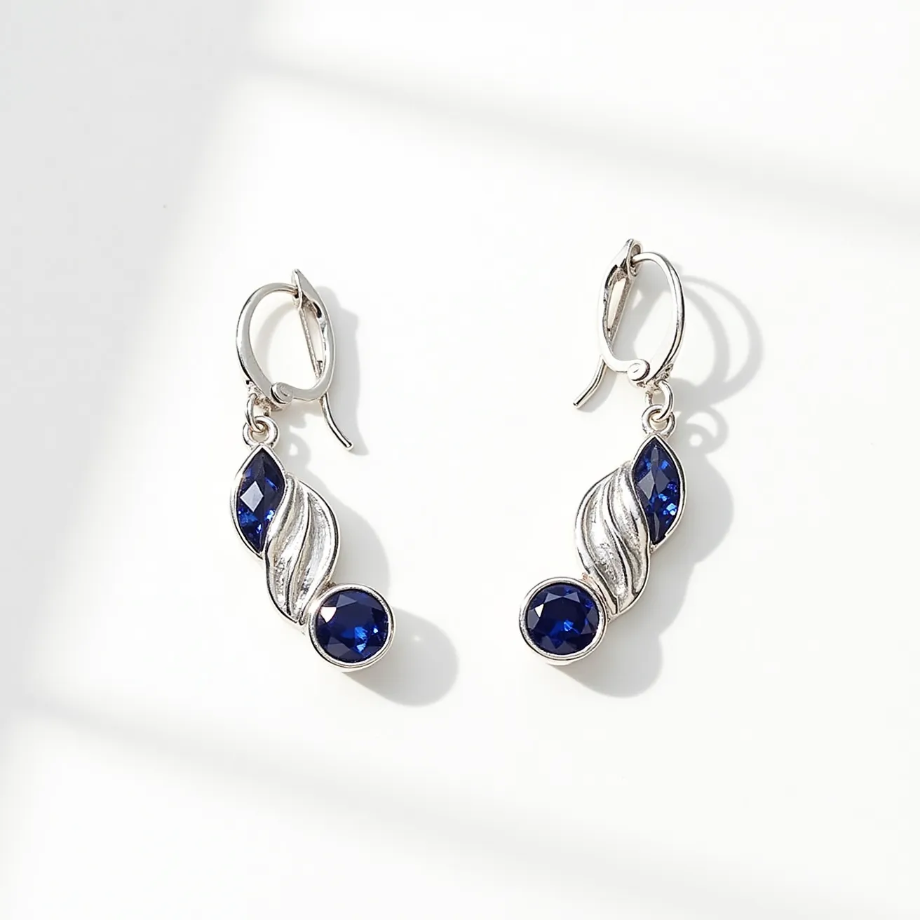 These silver earrings feature a graceful design adorned with deep blue gemstones. Each earring showcases two round-cut, faceted stones set in bezel settings, giving them a secure and polished appearance. Intertwined with these stones are elegant, leaf-like silver elements that add a flowing, organic aesthetic. The earrings are equipped with lever-back clasps, ensuring a secure and comfortable fit for the wearer. The combination of the rich blue gems and the sleek silver metal creates a classic yet contemporary look suitable for various occasions.