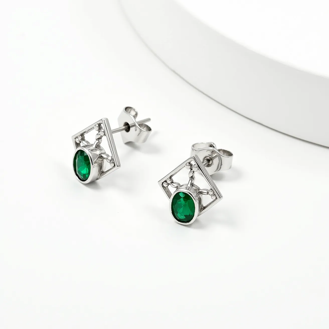 These silver earrings feature an elegant design with a prominent green gemstone at the center. The stones are oval-cut and are set within a rectangular frame, which adds a geometric appeal to the overall design. The setting showcases a prong or claw style, securely holding the gemstones in place while allowing light to enhance their verdant hue. The earrings are designed as stud earrings with a push-back clasp, ensuring ease of wear and security. The silver material gives the earrings a sleek and polished finish, complementing the richness of the green stones.