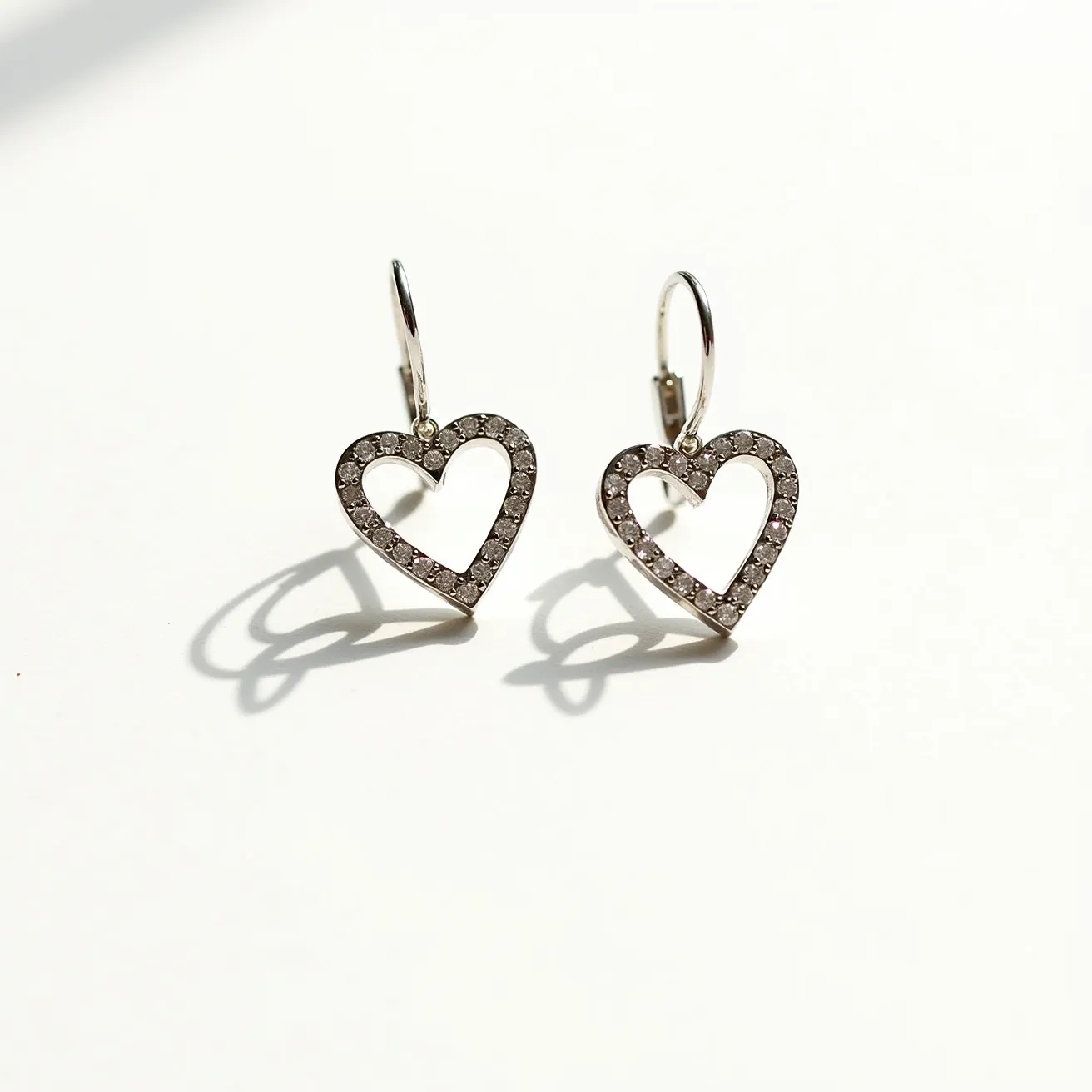 These silver heart earrings are crafted from a sleek and polished silver material, forming an elegant heart shape. They are adorned with small, round-cut stones set in a pavé setting, enhancing the shimmering effect throughout the design. The earrings feature a hook clasp for secure and comfortable wearing, allowing them to dangle gracefully. The meticulous arrangement of the stones along the heart’s outline adds a touch of sparkle, highlighting the craftsmanship of these delicate earrings.