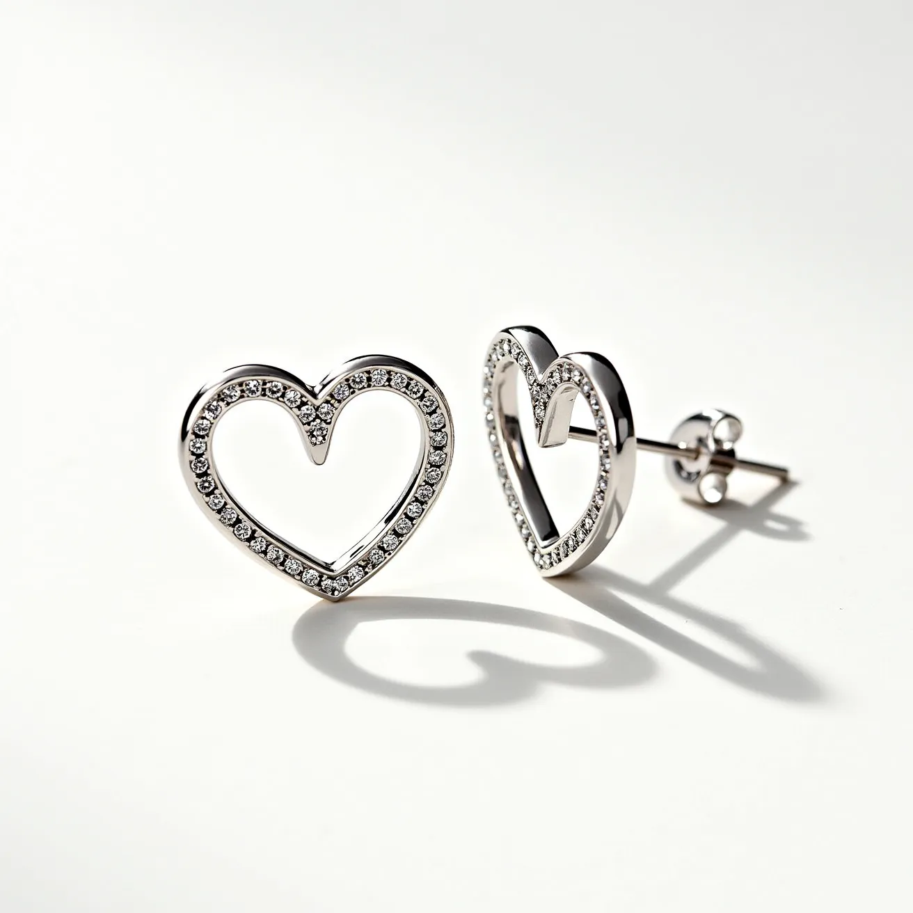 These silver heart earrings feature a delicate, open-work heart design crafted from polished silver. They are embellished with a row of small, round-cut gemstones, likely clear and sparkling, set along the front-facing edge of each heart, enhancing the elegance of the design. The stones are neatly set in a pave style, giving the earrings a continuous shimmer. The earrings use a post-back closure, suitable for pierced ears, providing secure and comfortable wear. These elements combine to create an elegant and timeless accessory.