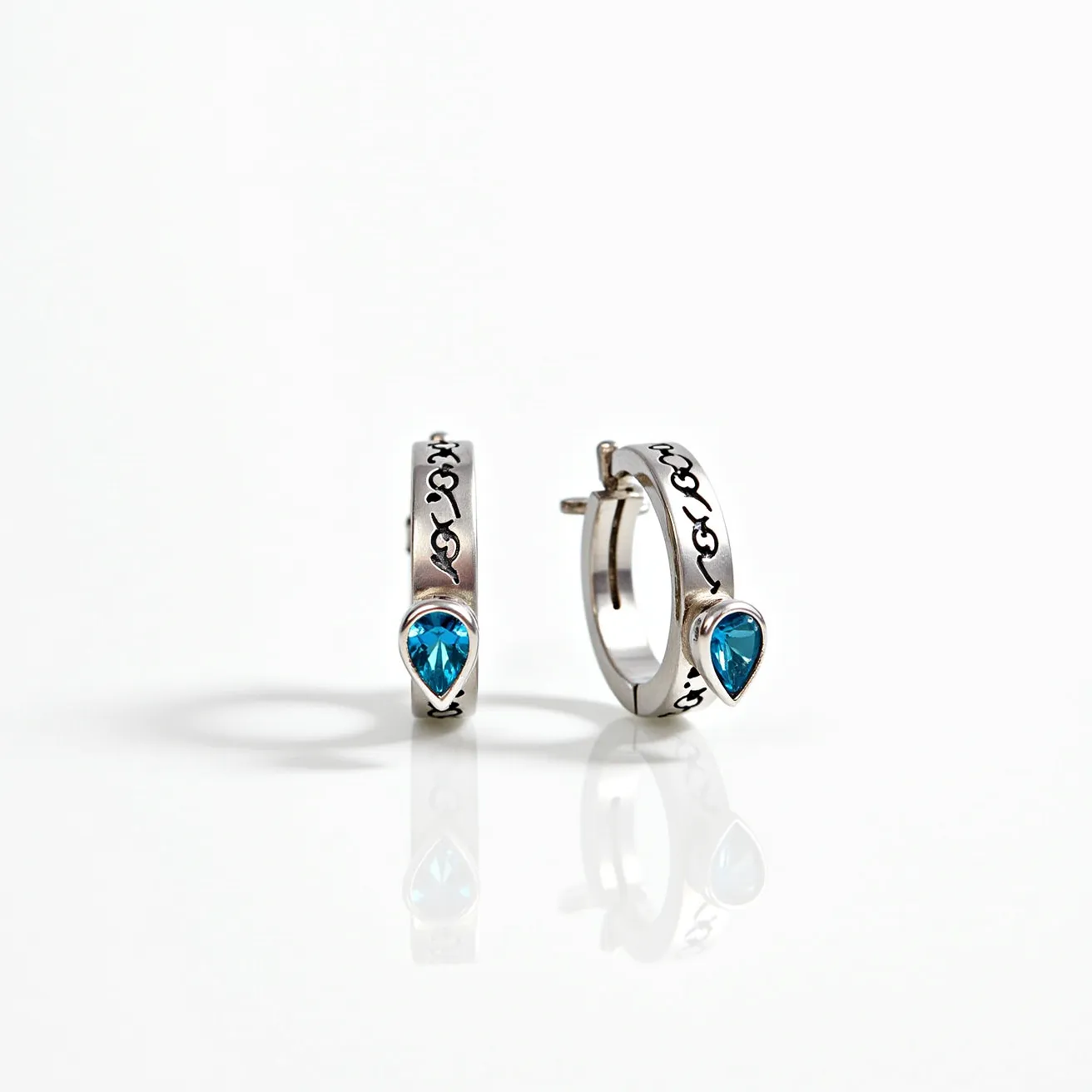 These silver hoop earrings feature a sleek design crafted from polished silver. They include a striking blue gemstone, cut in a teardrop shape, set securely within a bezel setting at the center of the hoop. The earrings are embellished with an intricate black pattern along the surface, adding an artistic touch. For attachment, they utilize a post-back clasp, ensuring a secure and comfortable fit.