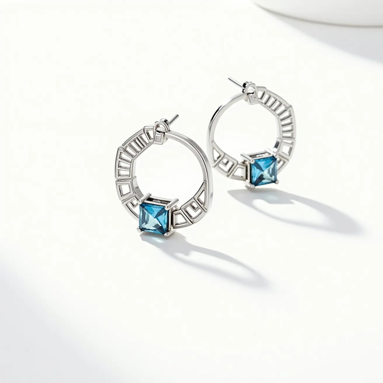 These silver hoop earrings feature an intricate design with square-cut blue gemstones as the centerpiece. The stones are securely held in a prong setting, enhancing their brilliance and visibility. The hoops are crafted from a polished silver material, which adds a sophisticated shine and complements the vibrant hue of the gems. The earrings include a post-back clasp for secure attachment, making them both comfortable and stylish to wear. The incorporation of geometric patterns with open spaces around the perimeter adds a modern touch to the overall design.