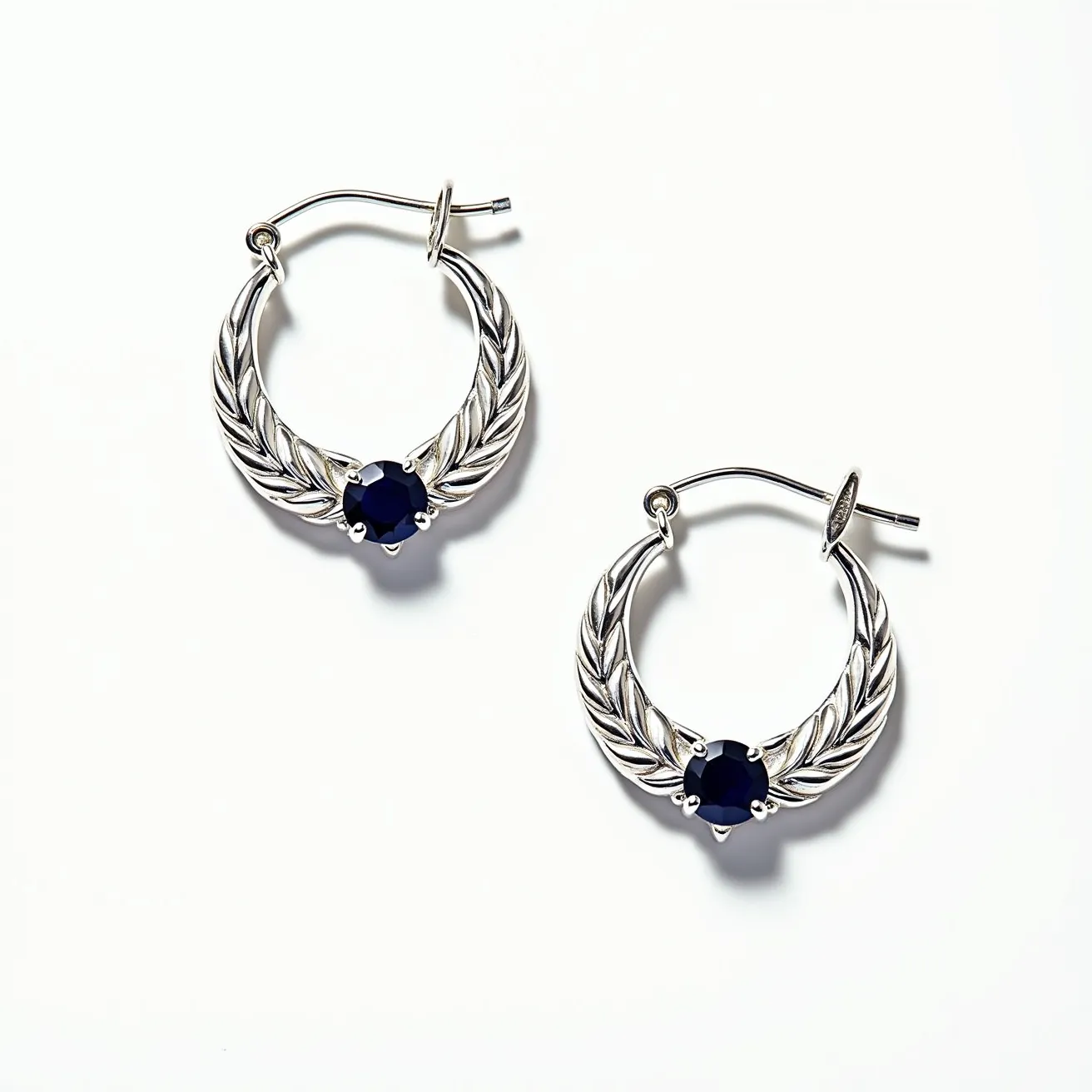 These silver hoop earrings are crafted with detailed feather-like engravings, giving them a sophisticated and unique look. At the center of each hoop is a deep blue, round-cut gem set in a prong setting, adding a touch of elegance and contrast against the silver tones. The earrings feature a latch back clasp, providing secure and comfortable wear. The combination of the intricate design and the striking blue gemstones makes these earrings a standout accessory.