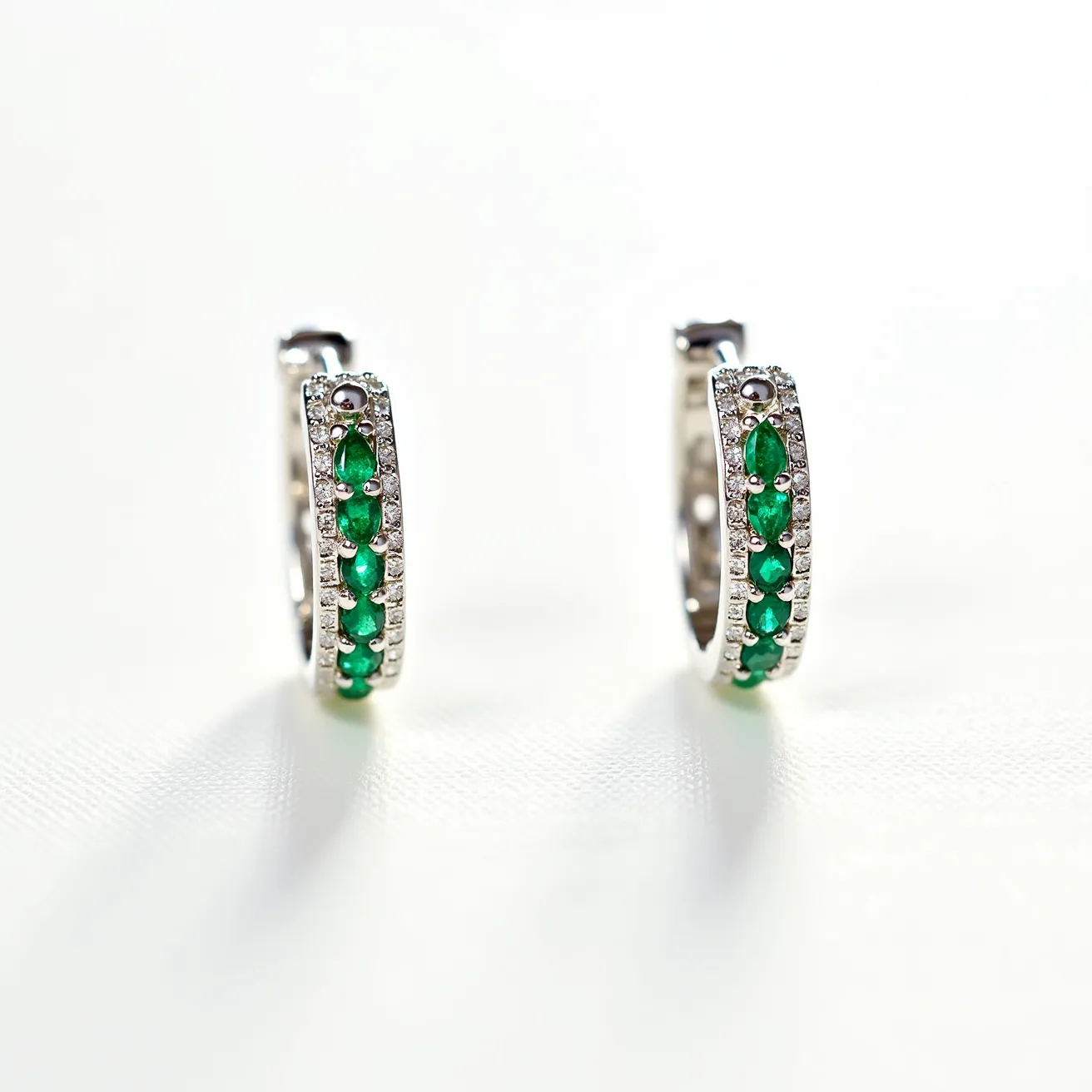 These silver hoop earrings feature an elegant combination of precious materials, with the main hoop crafted from polished silver. The front of each earring is adorned with small round green gemstones, likely emeralds, set in a neat row. These stones are bordered by a line of sparkling white stones, possibly diamonds or cubic zirconia, all set in a pave setting that enhances their brilliance. The earrings are secured with a hinge-back clasp, ensuring both style and ease of use. The overall design is refined, with a modern touch brought by the vibrant green and delicate white stones.