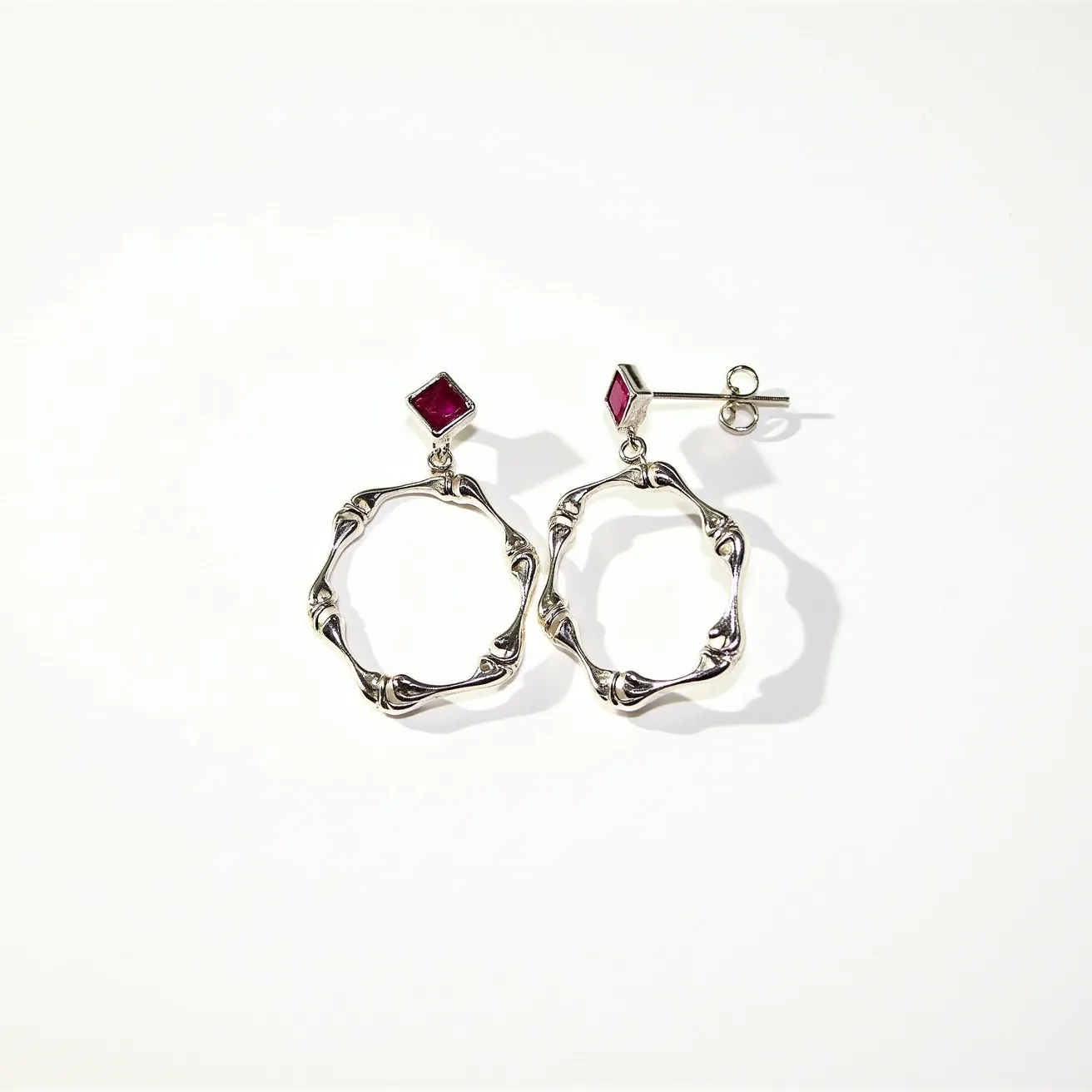 These silver hoop earrings feature a dynamic and artistic design, made from a high-quality silver metal. Each earring is adorned with a square-cut red gemstone, likely a ruby or garnet, set in a bezel setting just above the hoop, adding a touch of elegance and color contrast. The earrings are designed with a post and butterfly clutch fastening, ensuring a secure and comfortable attachment. The hoops themselves have an organic, wavy form that adds to the unique and modern aesthetic of the piece.