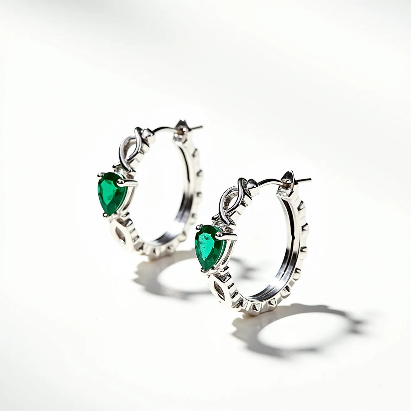 These silver hoop earrings are crafted from a shiny silver material with a polished finish. Each earring features a vibrant green gemstone with a pear cut, securely set in a three-prong setting that adds elegance and color contrast to the overall design. The hoops have a ridge pattern along the edges, enhancing their visual interest. For attachment, the earrings utilize a post-and-hinge clasp mechanism, providing a secure and comfortable fit for the wearer.