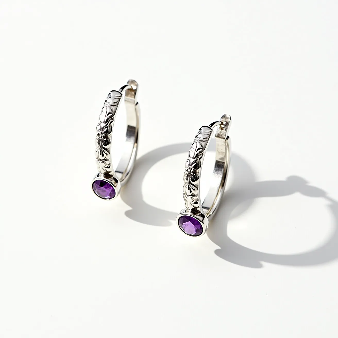 These silver hoop earrings feature a classic design with detailed floral engravings on the outer surface, adding an elegant and intricate touch. Each earring incorporates a round, faceted amethyst gemstone, set in a bezel setting at the base of the hoop. The vibrant purple hues of the gemstone contrast beautifully with the polished silver, enhancing its visual appeal. These earrings have a lever-back clasp, providing a secure and stylish fastening. The craftsmanship combines traditional elegance with a modern twist, making these earrings a versatile accessory for any occasion.