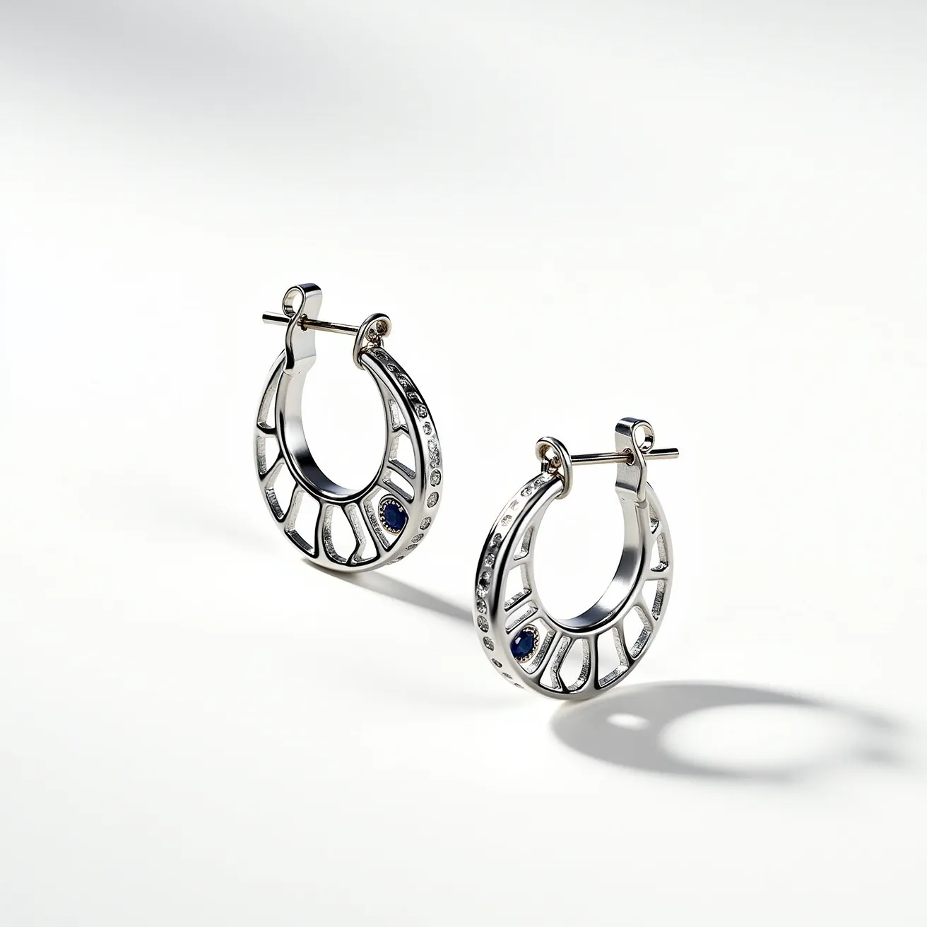 These silver hoop earrings feature a sleek, polished metal design, possibly sterling silver, with a series of inset clear gemstones lining the outer edge, adding a subtle sparkle. At the bottom of each hoop is a single, larger blue gemstone, likely a sapphire, set flush within the metal for a modern, cohesive look. The earrings are designed with a hinge clasp mechanism at the top, which secures the hoop effectively in place when worn. The overall design combines classic elegance with contemporary flair, making them a versatile accessory.