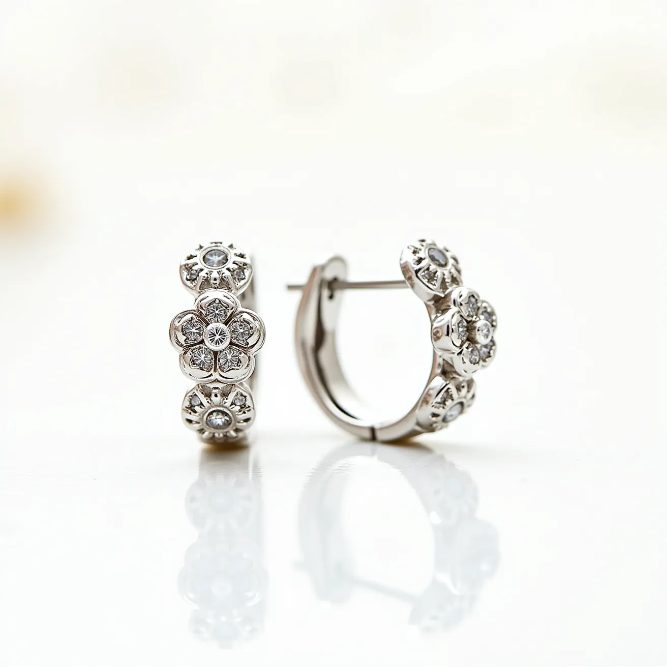 These silver huggie earrings feature intricate floral designs adorned with multiple clear, round-cut gemstones, which resemble small diamonds or cubic zirconia. Each earring showcases a row of flower motifs, meticulously crafted to enhance their elegance. The stones are encased securely in a bezel setting, which not only holds them in place but also adds a sleek, modern touch to the pieces. The earrings utilize a hinged clasp mechanism, ensuring a secure and comfortable fit against the earlobe, characteristic of huggie-style designs. The polished silver finish gives the earrings a sophisticated and timeless appeal.