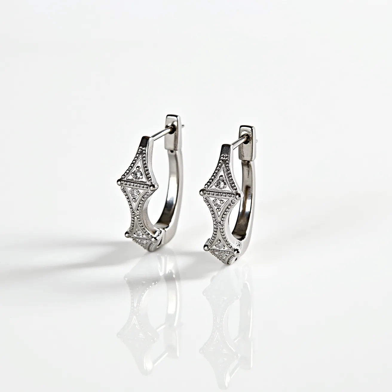 These silver huggie earrings feature an intricate design with an elegant, geometric pattern adorned with small, clear stones. The stones are likely set in a pavé or micro-pavé setting, adding a refined sparkle to the earrings. The main structure appears to be crafted from sterling silver, providing a sleek and polished look. They secure with a hinged backing, characteristic of huggie earrings, ensuring a snug fit on the earlobe. The overall craftsmanship combines modern style with a subtle vintage touch through the detailed embellishments.