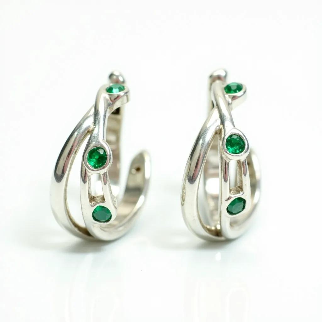 These silver huggie earrings feature a sleek and polished design, crafted from a high-quality silver material that offers a sophisticated shine. They are adorned with round-cut green gemstones, possibly emeralds, which are securely set into the earrings with a bezel setting that enhances their vibrant color and adds a touch of elegance. The earrings use a hinged clasp mechanism, typical of huggie styles, ensuring a snug and comfortable fit around the earlobe. These elements come together to create a piece that is both stylish and versatile, suitable for various occasions.