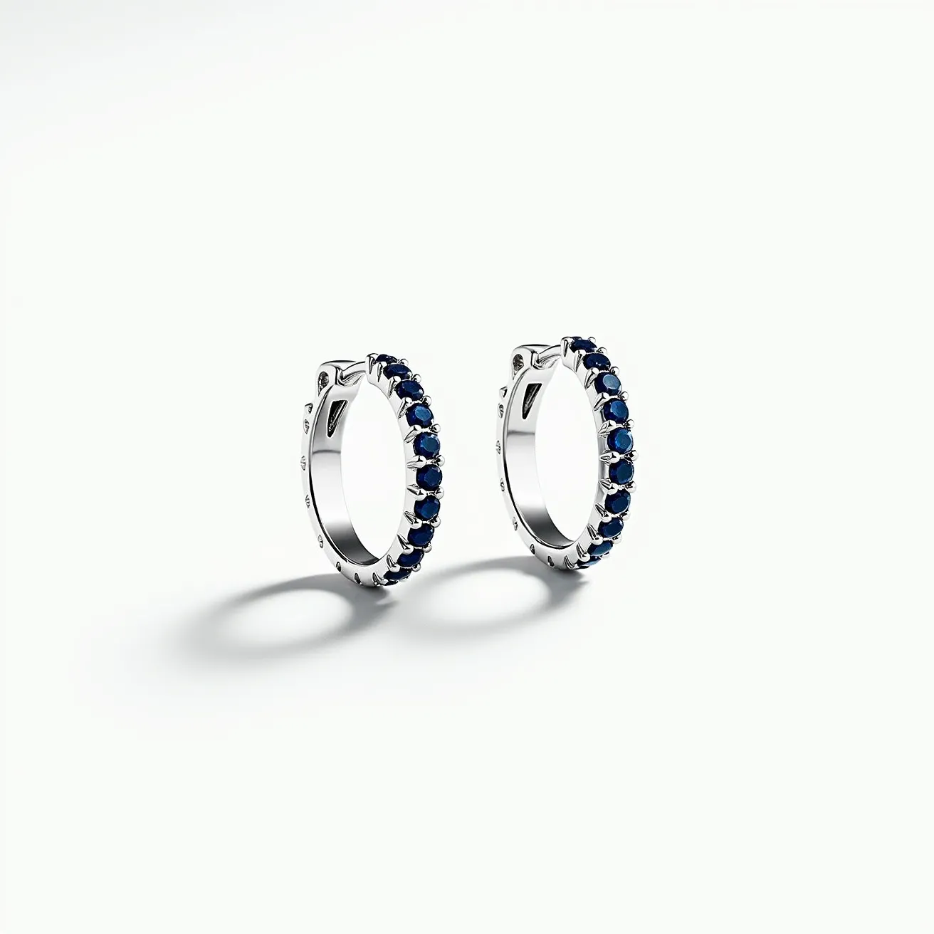 These silver huggie earrings feature a sleek design crafted from silver, boasting a polished finish. They are embellished with a row of blue gemstones, likely sapphires, each cut into small, uniform round shapes. The stones are securely set in a prong setting, allowing them to catch the light beautifully. The earrings are designed with a hinged clasp mechanism, ensuring they fasten snugly to the earlobe, providing both comfort and security for everyday wear.