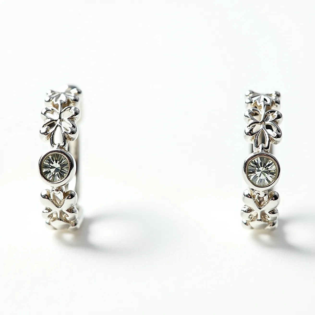 These silver huggie earrings feature a delicate design, crafted from high-quality silver. They incorporate intricately shaped motifs that resemble floral or leaf patterns. Each earring is adorned with a single round gemstone, which is set in a bezel setting, adding a touch of elegance and sparkle. The gemstones, possessing a clear appearance, are finely cut to reflect light. The earrings utilize a hinge and clasp mechanism, allowing for secure and comfortable wear.