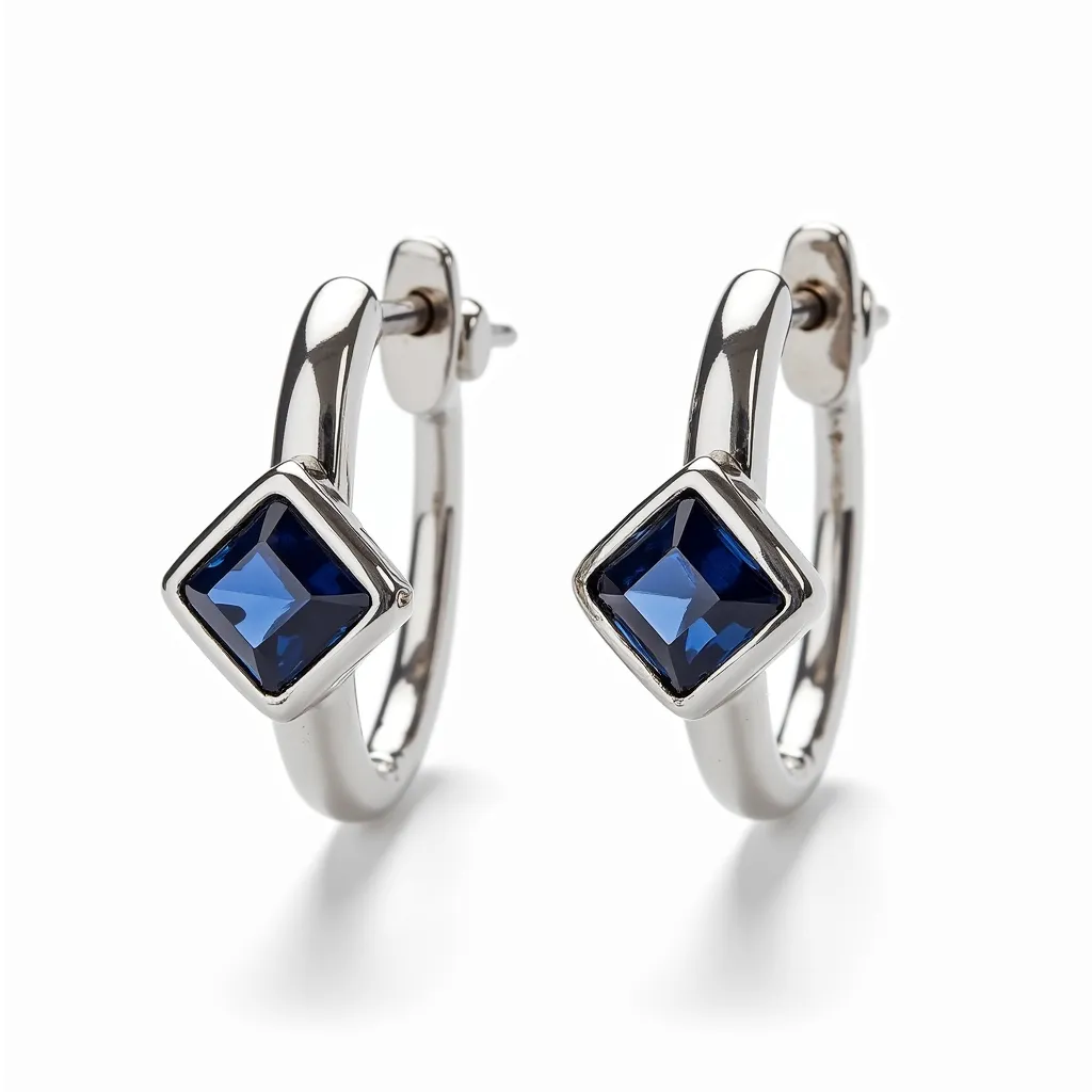 These silver huggie earrings are crafted from a refined silver metal, showcasing a sophisticated design. Each earring features a striking square-cut blue gemstone, held securely in a bezel setting, which beautifully complements the sleek metallic finish. The huggie style offers a comfortable fit with its hinged clasp mechanism, ensuring the earrings remain securely in place when worn. The combination of the rich blue gemstone and the gleaming silver metal creates a contemporary yet elegant aesthetic, suitable for various occasions.