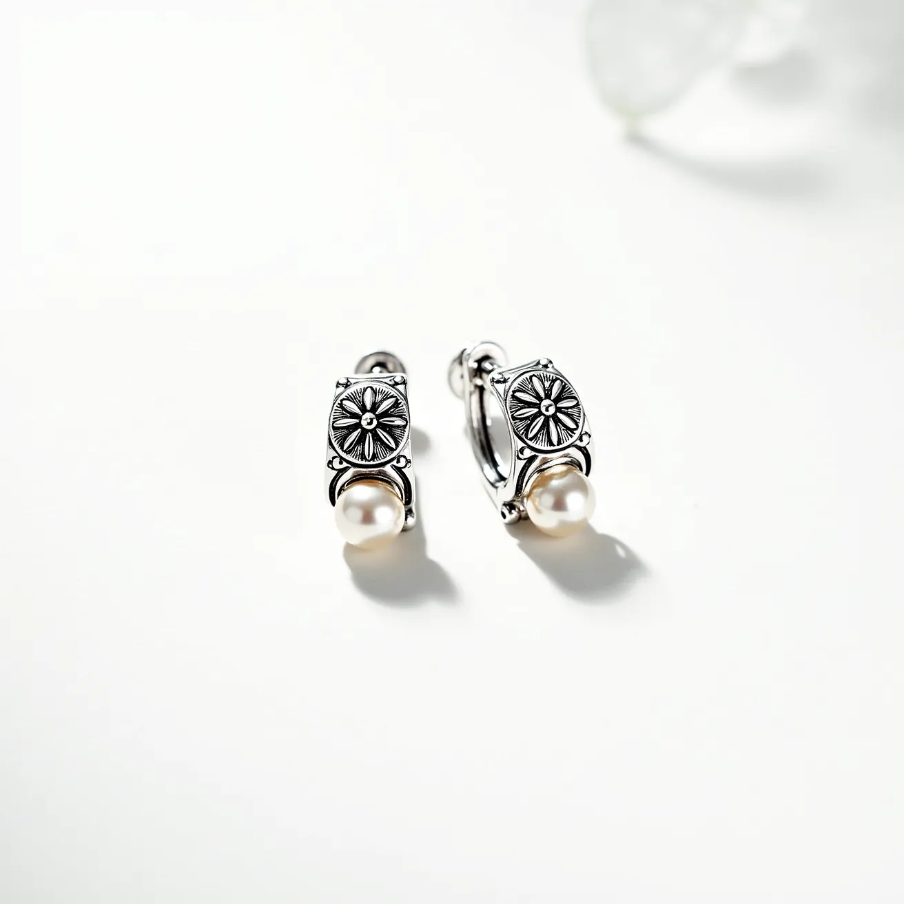 These silver huggie earrings feature a polished metal design with an intricate flower motif engraved into the outer surface, adding a touch of elegance and intricate craftsmanship. Each earring is adorned with a lustrous pearl set at the base, accentuating the overall sophistication with its smooth and reflective surface. The earrings are designed with a classic huggie clasp, ensuring a secure and comfortable fit for everyday wear. The combination of the silver metal and the pearls creates a timeless aesthetic that is both versatile and stylish.