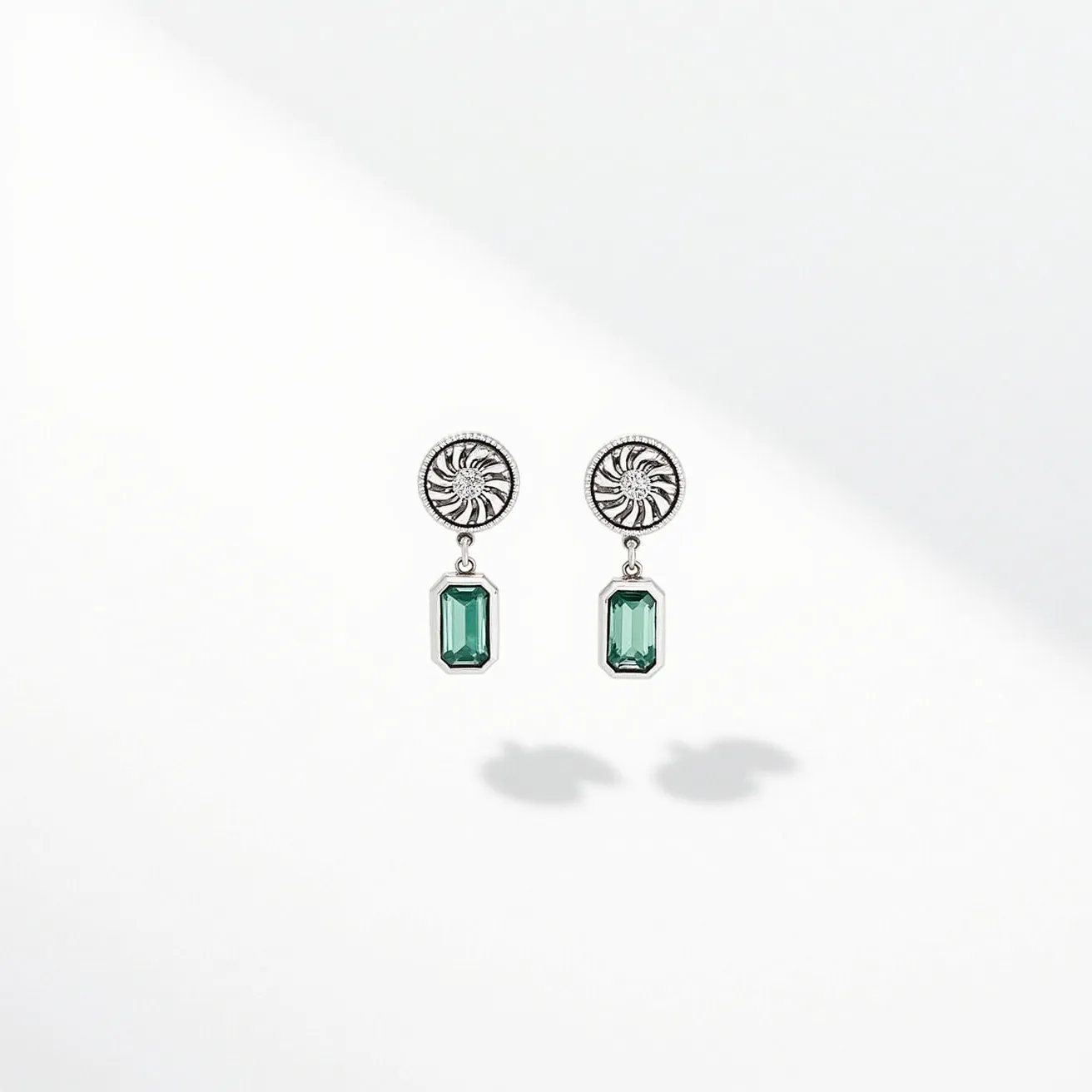 These silver stud earrings feature a stylish design with a circular stud embellished with a swirling pattern. Suspended from each stud is a rectangular green gemstone, showcasing an emerald cut that enhances its reflective surface and depth. The gemstones are held in place with a prong setting, allowing maximum light to pass through and highlight their vibrant color. These earrings are designed with a post back, providing a secure and comfortable fit for the wearer.