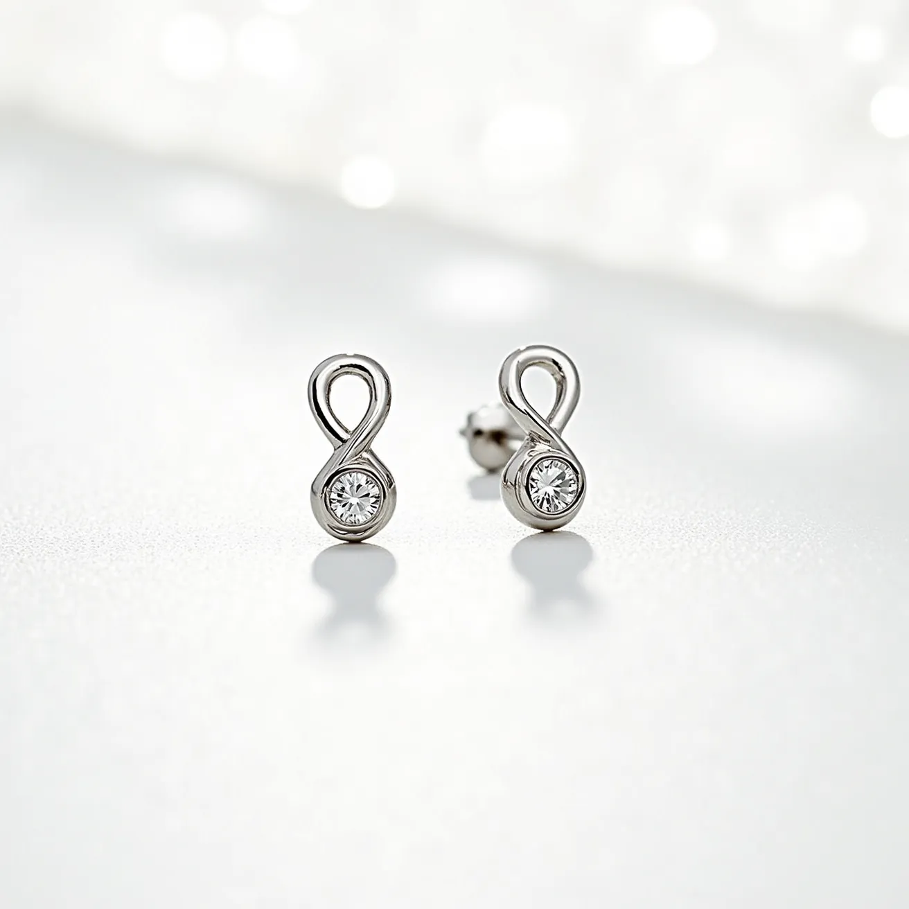 These silver stud earrings feature a sleek, elegant design, crafted primarily from polished silver shaped into an infinity symbol. At the base of each earring is a round, clear gemstone, likely a diamond or cubic zirconia, set in a bezel setting which securely encases the stone. The stones are faceted, enhancing their brilliance and allowing them to catch light effectively. The earrings are secured with a classic post and clutch back fastening, ensuring a snug fit when worn. These carefully designed elements come together to create a timeless and sophisticated accessory.