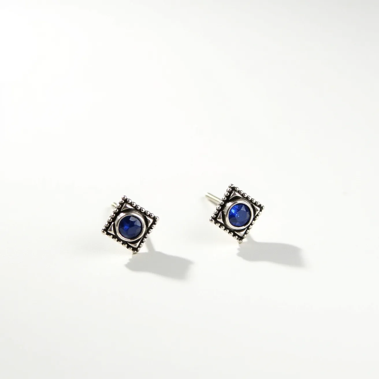 These silver stud earrings feature a central round blue gemstone, possibly a sapphire or a similar stone, prominently set in a bezel setting. The gemstone is surrounded by a square silver frame embellished with small beaded details that add texture and dimension to the design. The earrings have a classic post attachment, ensuring secure wear. The overall look combines elegant simplicity with intricate detailing, making them a versatile accessory.