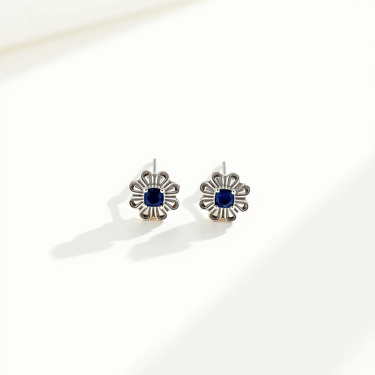 These silver stud earrings feature a floral-like design crafted from silver, accentuated by a central blue gemstone. The gemstone appears to be set in a secure prong setting, adding a vibrant contrast to the silver petals surrounding it. The cut of the gemstone is a square shape with facets that enhance its deep, rich color. The earrings are attached with a post back, which is a common type of clasp for stud earrings, ensuring easy and comfortable wear.