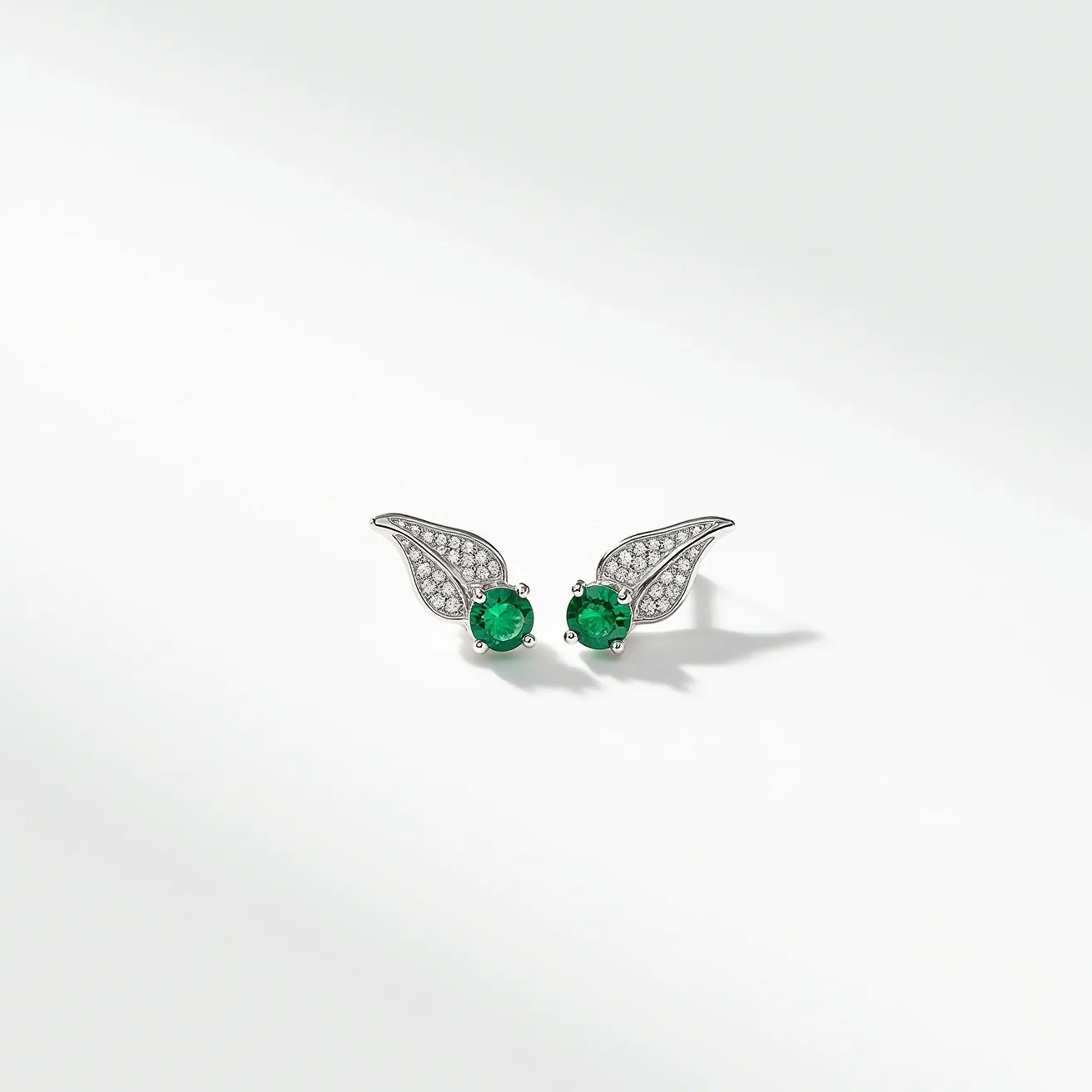 These small earrings feature a striking design with central green gemstones, likely emeralds, each exhibiting a round cut to enhance their brilliance. Surrounding the emeralds, the setting includes what appear to be clear, small round stones, possibly diamonds, arranged artistically in a wing-like pattern. The metal used for the setting appears to be a white metal, potentially silver or white gold, providing an elegant backdrop to highlight the stones' colors. The earrings are designed as studs, suggesting a post and clutch back style of attachment, ensuring they are secure when worn.