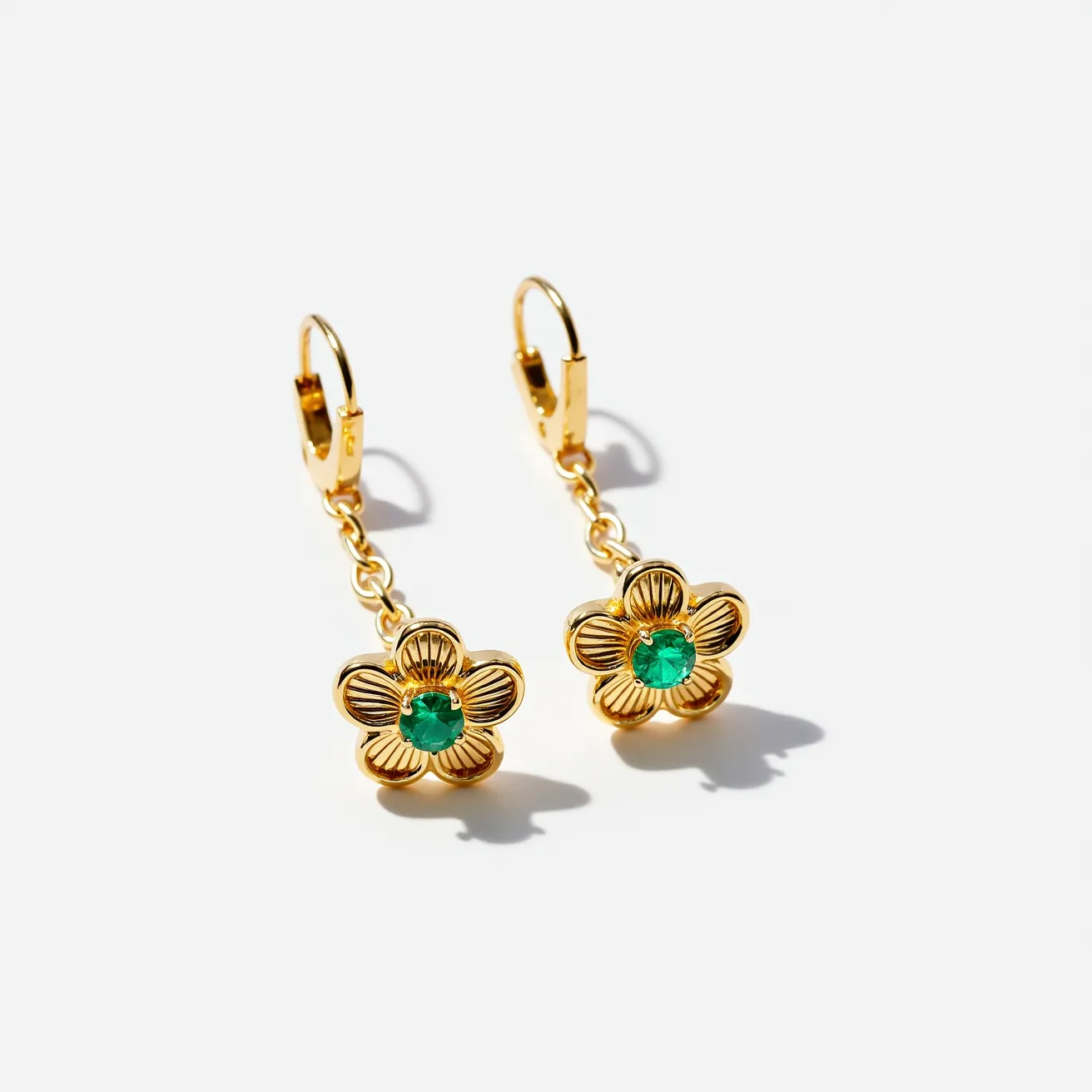 These small earrings feature a floral design crafted from a gold-toned metal, with a central green gemstone in each earring. The gemstones are likely emeralds, cut in a round shape and set in a bezel or prong setting. The flower petals have a textured appearance that adds depth and interest to the design. These earrings are attached to a short, delicate gold-toned chain which connects to a lever-back clasp, providing a secure closure and adding an elegant dangle effect to the overall composition.