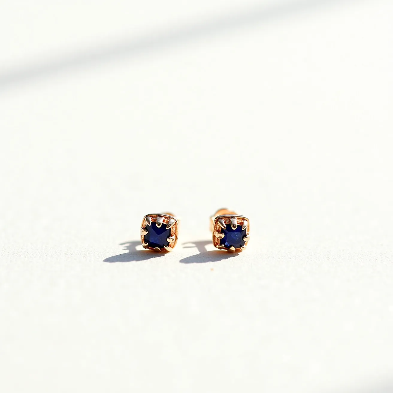 These small earrings feature a central gem with a deep blue color, cut in a square shape, and set in a prong setting. The prongs appear to be made of a gold-toned metal, adding a classic and elegant touch. The earrings likely use a post-back attachment, which is common for securing stud earrings comfortably. The combination of the blue gem with the gold setting creates a striking contrast that enhances the overall aesthetic of the earrings.