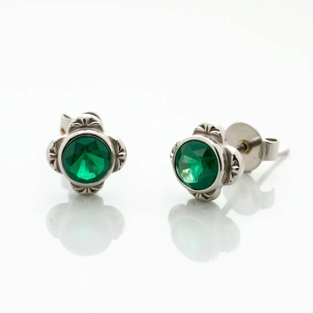 These small earrings feature a central green gem, likely an emerald or emerald-like stone, cut in a round shape and set in a decorative bezel setting. The metal appears to be silver or white gold, forming a floral or clover design around each stone that adds an elegant touch. The earrings are equipped with push-back clasps, securing the post that delicately supports the overall structure. The craftsmanship highlights both the vibrant stone and the intricate metalwork, creating a harmonious and refined accessory.