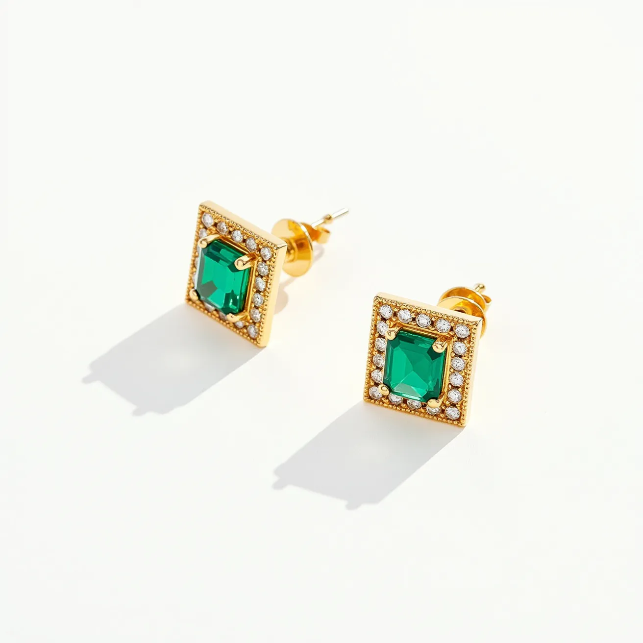 These square earrings feature a striking design with a central emerald-colored gemstone in a square cut, securely held in place by four prongs in a bezel setting. The gemstone is surrounded by a border of small, round, clear stones that add an extra layer of sparkle. These stones are set in a gold-toned metal, creating a luxurious frame around the centerpiece. The earrings attach via a classic post and butterfly clasp, ensuring they stay securely in place when worn.