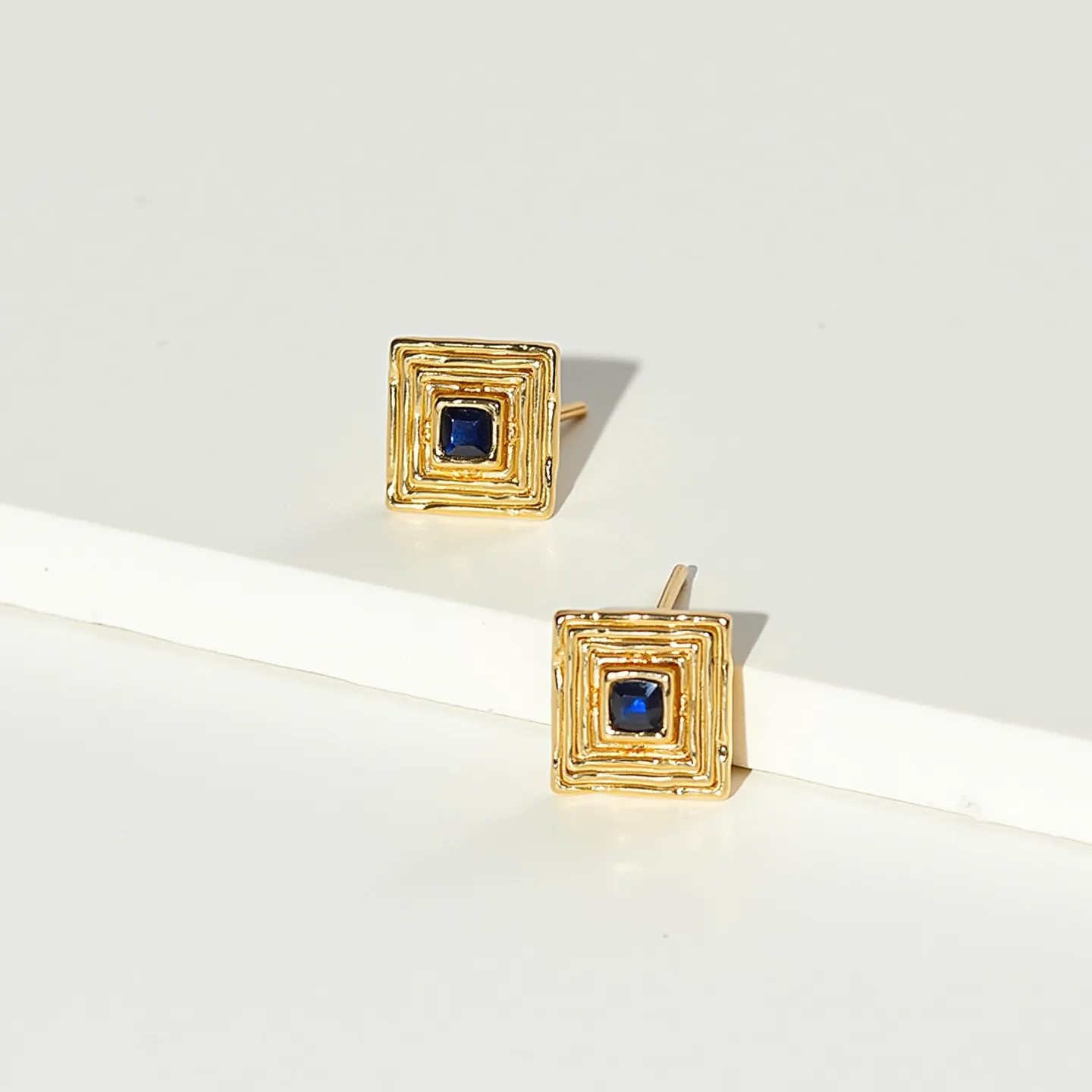 These square earrings feature a geometrically structured gold-tone metal design, characterized by concentric square layers that create a textured, visually appealing surface. At the center of each earring is a square-cut blue gemstone, likely a sapphire, set securely to highlight its deep color and sparkle. The earrings are equipped with straight posts, suitable for pierced ears, ensuring a simple attachment and elegant look. The combination of the gold-toned metal and blue gemstone provides a classic and sophisticated style.