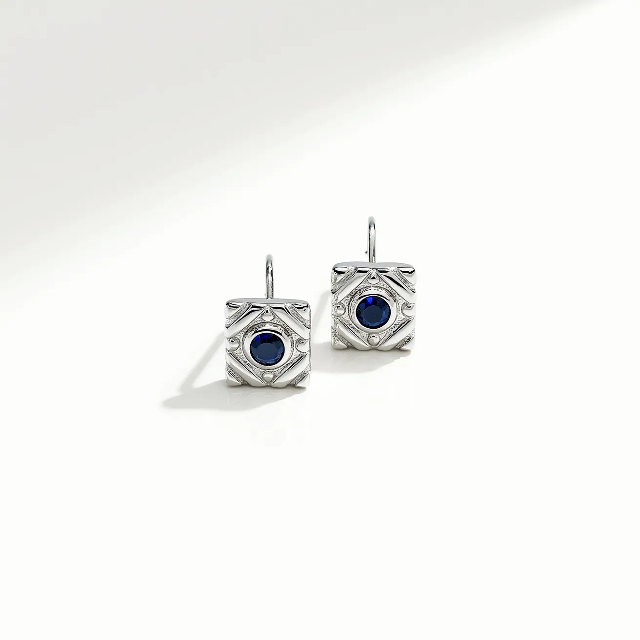 These square earrings feature a sleek and elegant design crafted from a silver-toned metal, giving them a sophisticated and timeless appearance. At the center of each earring is a round blue gemstone, likely a sapphire, that is bezel-set, providing a secure and polished look. The metal surface is intricately engraved with geometric patterns that add texture and depth to the overall design. The earrings are equipped with a simple hook attachment, making them easy to wear and offering a classic style that complements a variety of outfits.