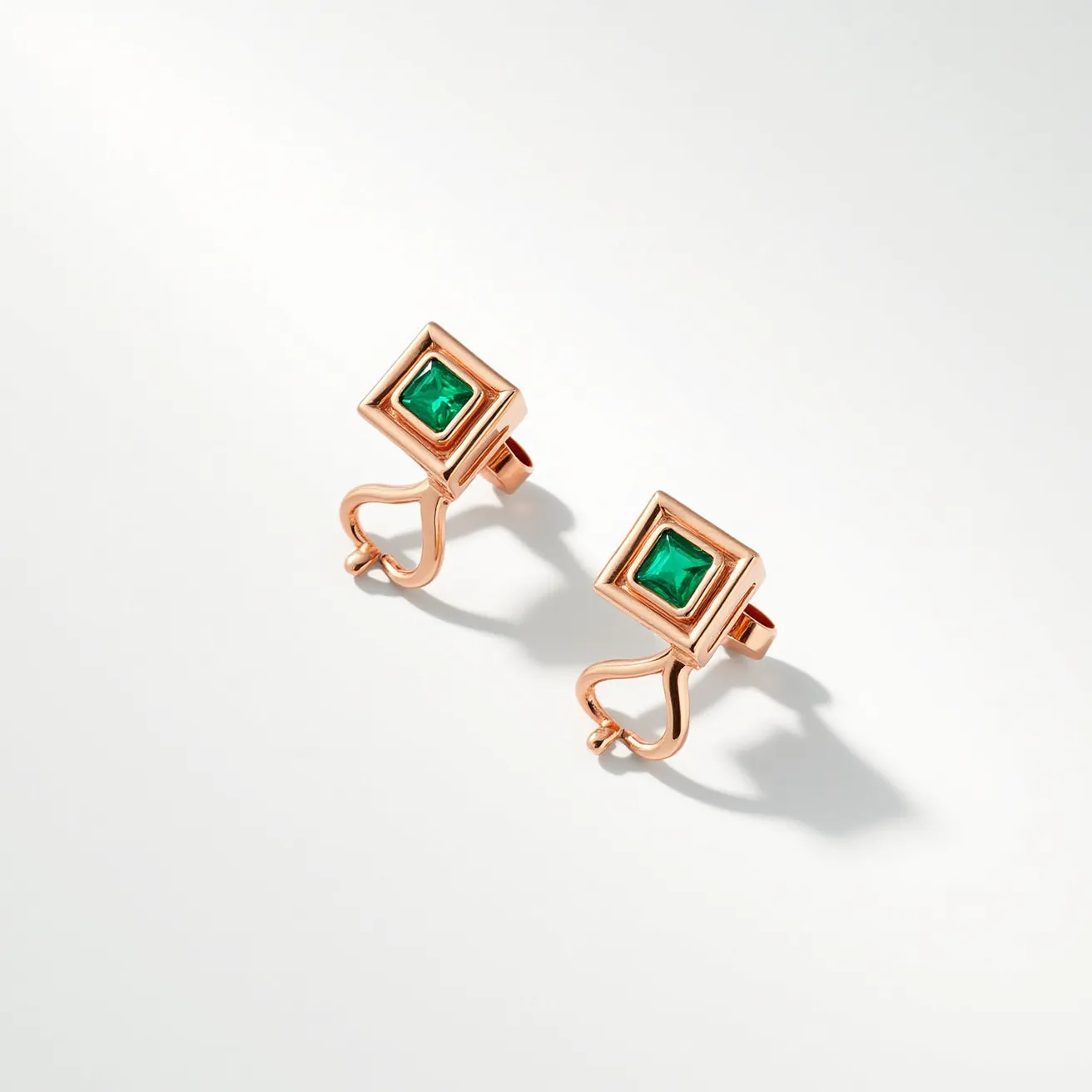 These square earrings feature a sophisticated design with a prominent square cut green gemstone set at the center of each piece. The gemstones are likely emeralds, known for their rich, captivating hue, and are securely held in place by a bezel setting that frames each stone with precision. The metallic structure of the earrings appears to be crafted from a rose gold material, adding a warm and elegant contrast to the vivid green of the gemstones. The earrings are attached to a unique metal framework that curves gracefully, ending in a post-back clasp for secure fastening. The overall design combines modern geometric elements with traditional gemstone settings, creating a versatile and stylish accessory.