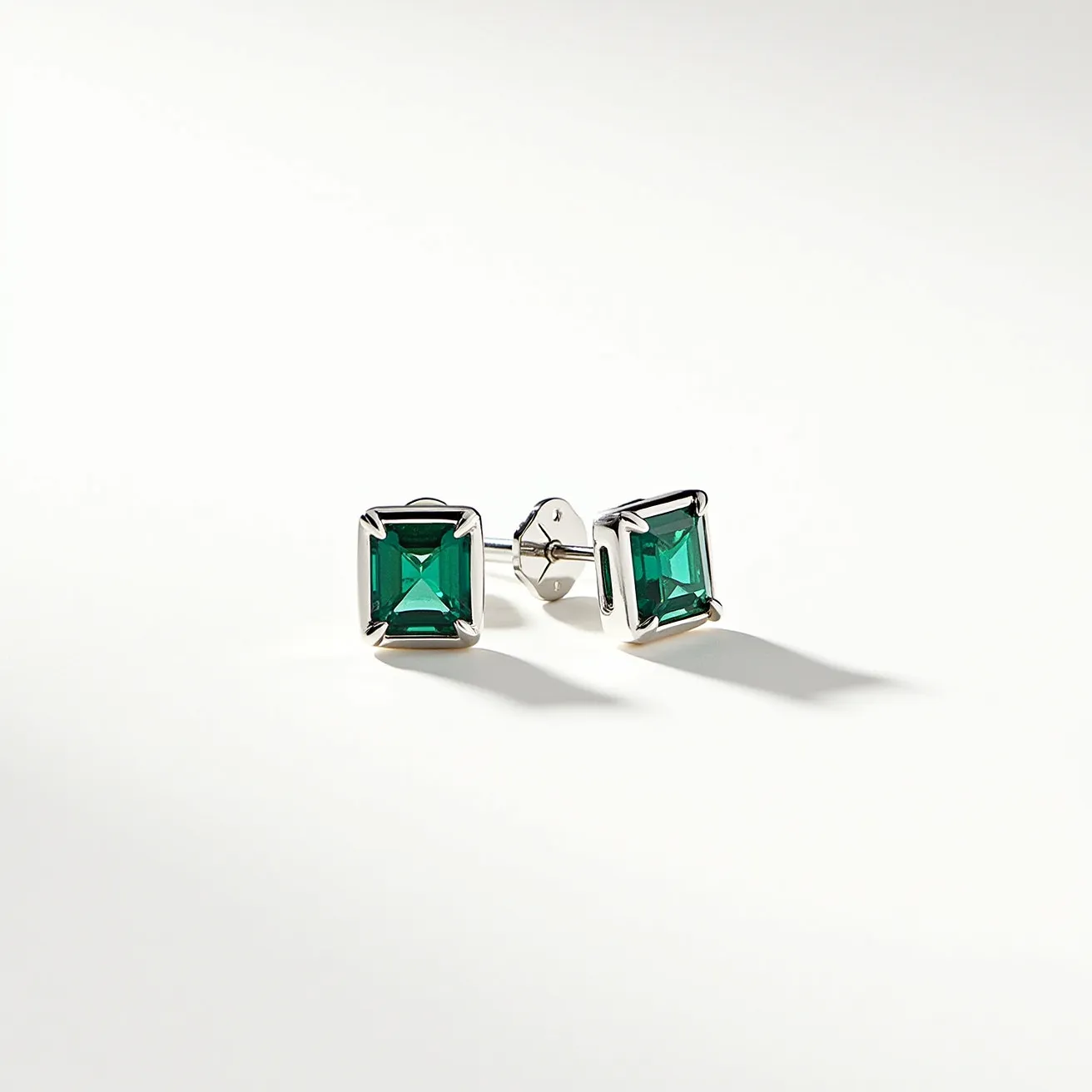 These square earrings feature a vibrant green gemstone, cut in a square shape and set in a sleek, polished metal that may likely be silver or white gold, enhancing their elegance. The gemstones are held securely in place by a four-prong setting, subtly showcasing their clarity and richness. The earrings are equipped with a post-back clasp, providing a secure attachment while wearing. The overall design is minimalistic yet sophisticated, allowing the beauty of the gemstones to stand out prominently.